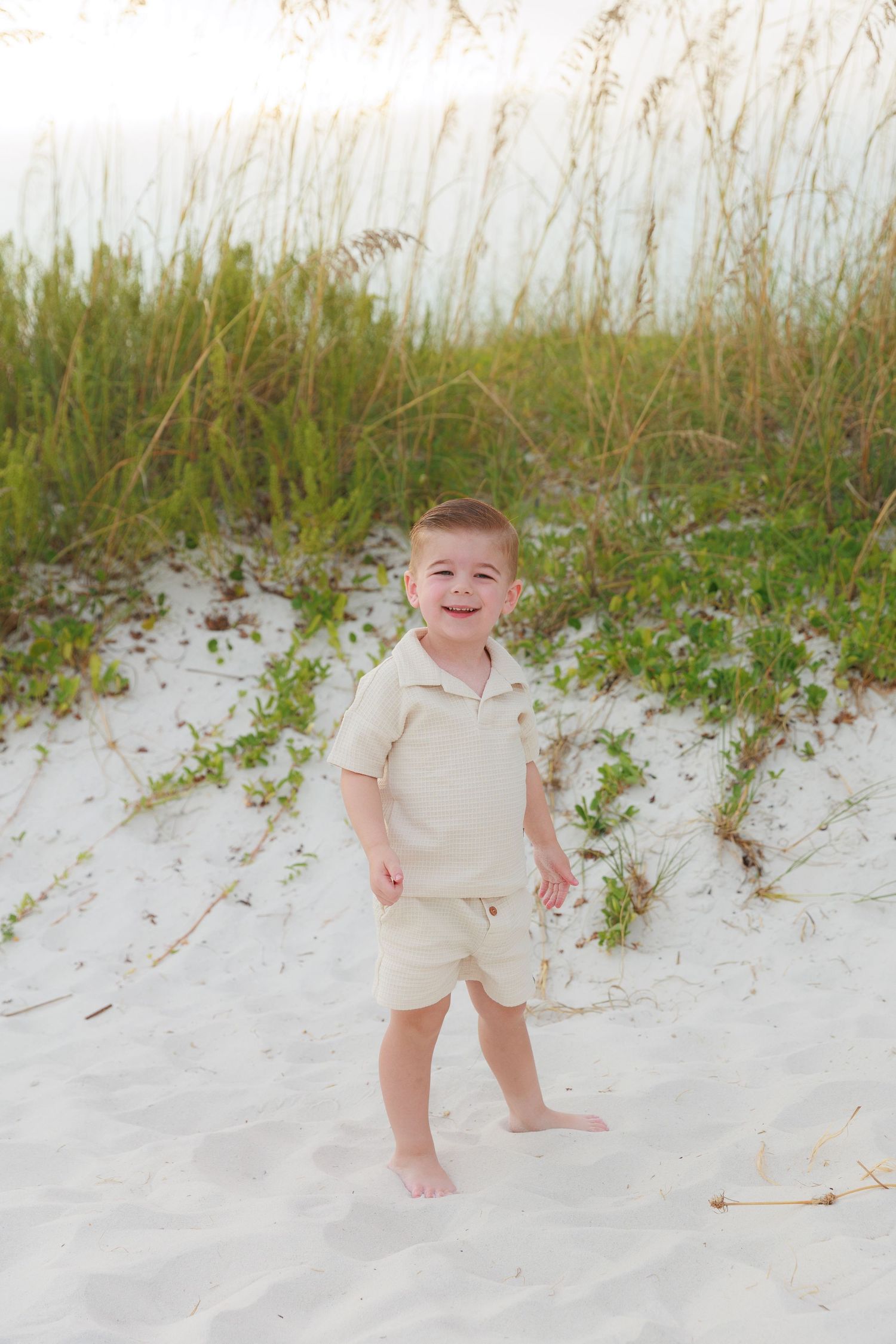 Happy toddler boy in beige outfit smiling on white sand beach dunes with sea oats grass background.