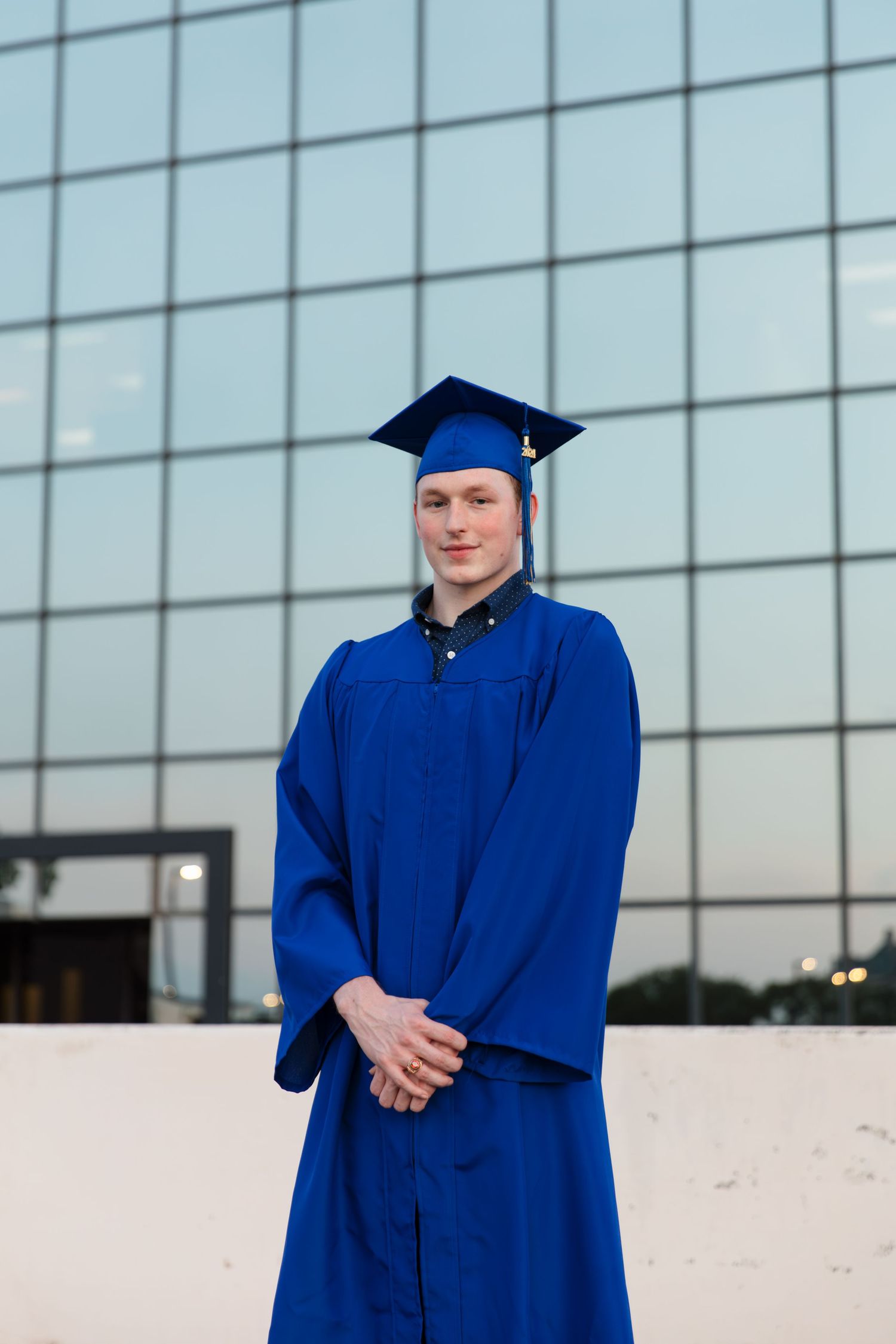 Young man in blue graduation cap and gown standing confidently outside a modern glass building.