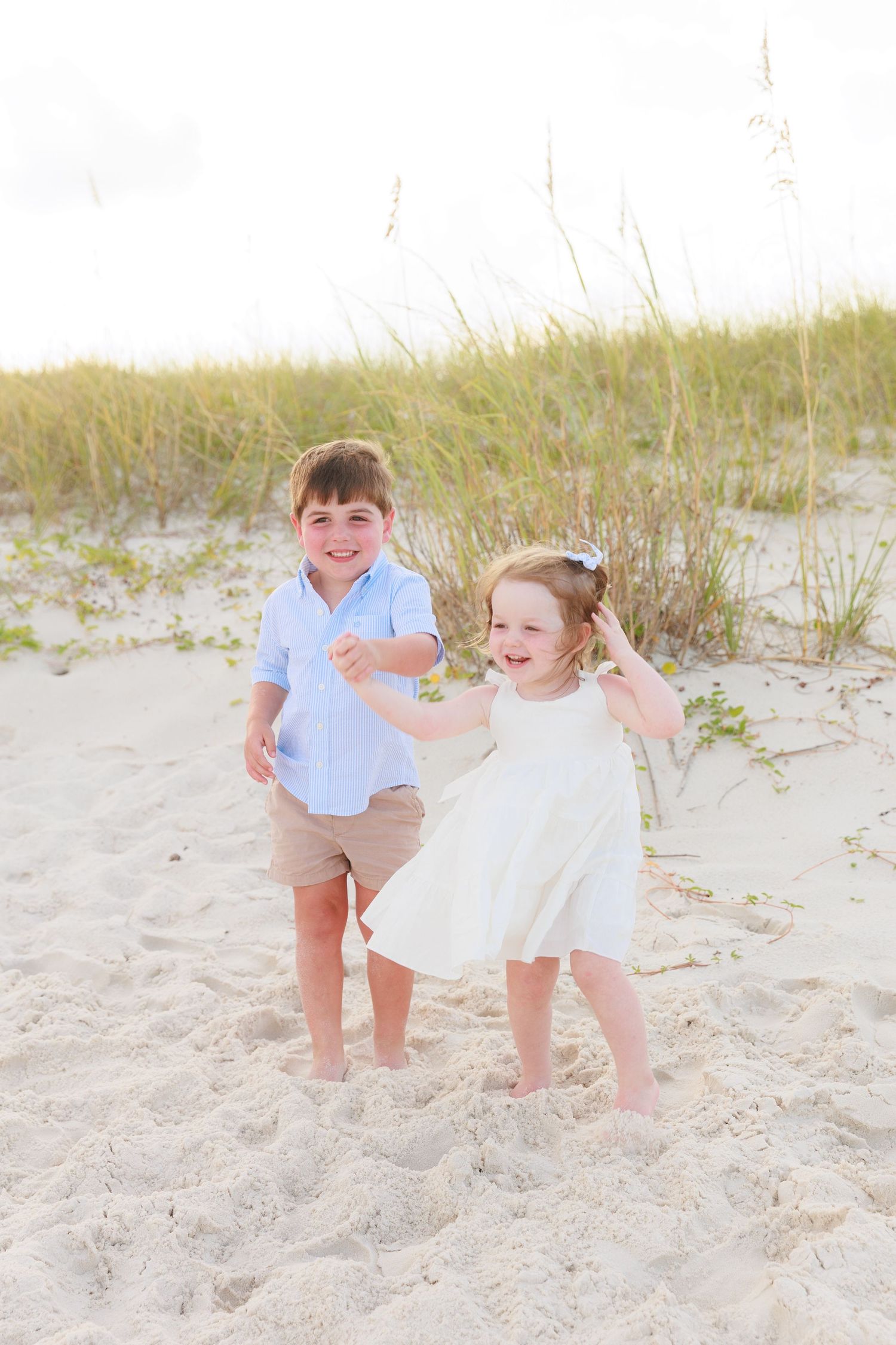 Two happy children playing on a sandy beach near sea oats, boy in blue shirt and girl in white dress.