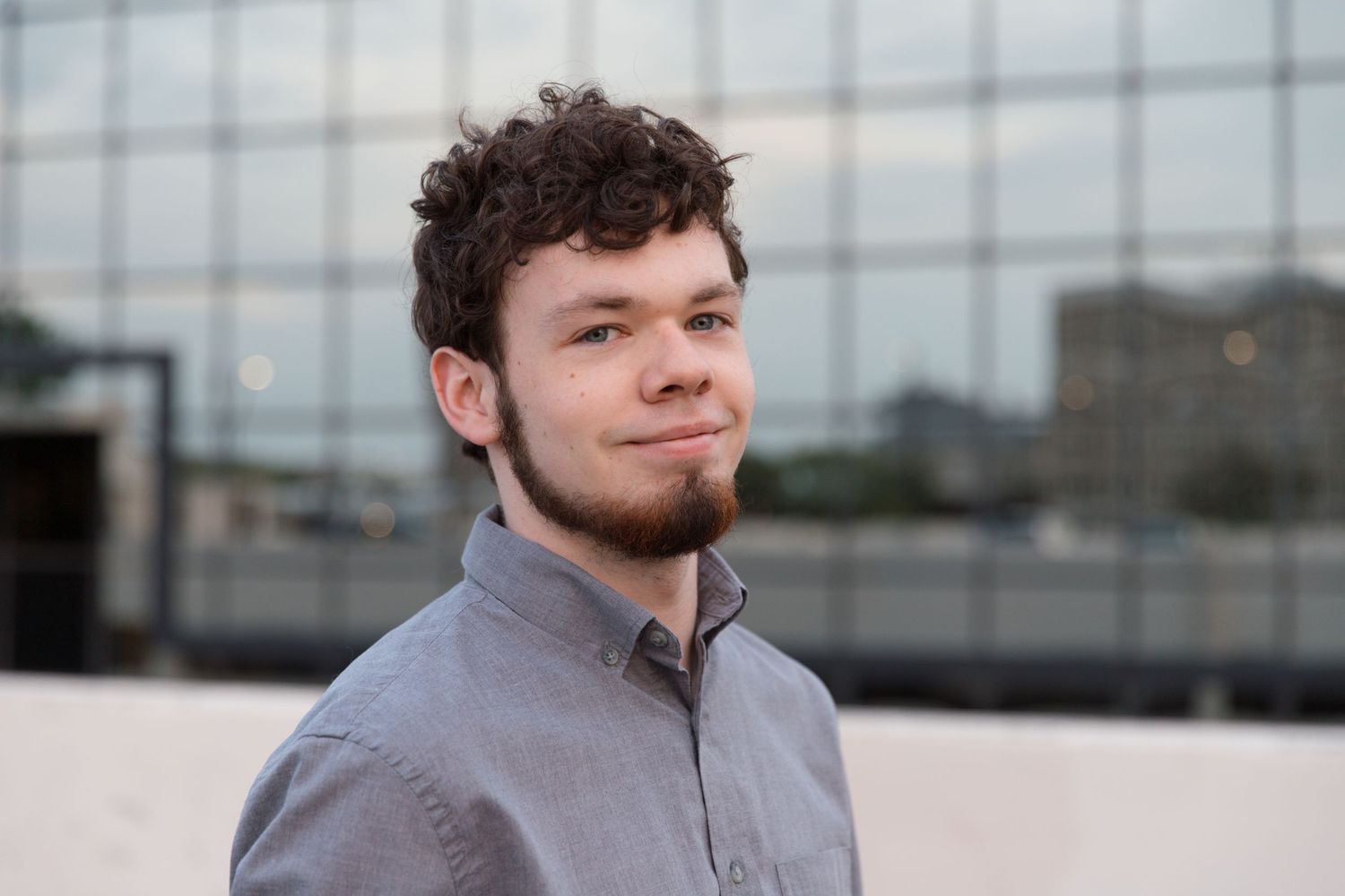 Professional headshot with glass building backdrop wearing grey collared shirt.