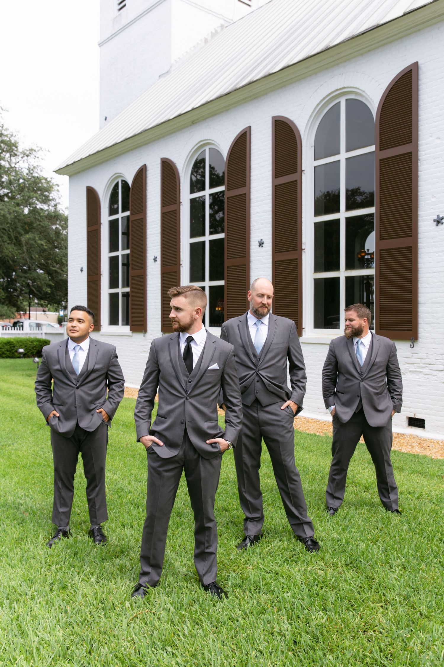 Groomsmen in gray suits pose casually in front of white church with arched windows.
