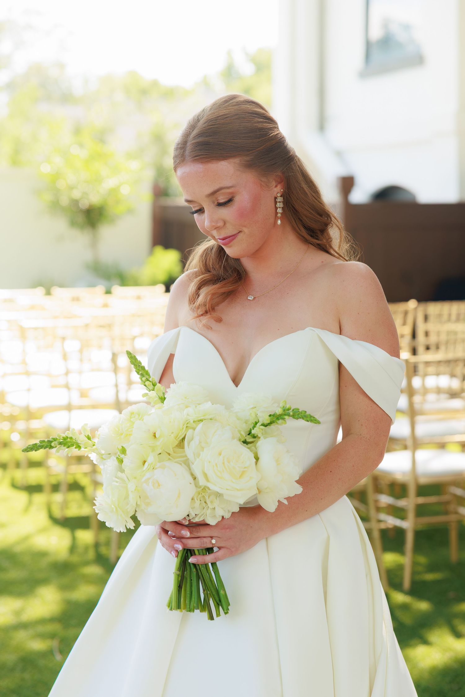 Bride in off-shoulder white ballgown holding white peony bouquet outdoors at wedding venue.