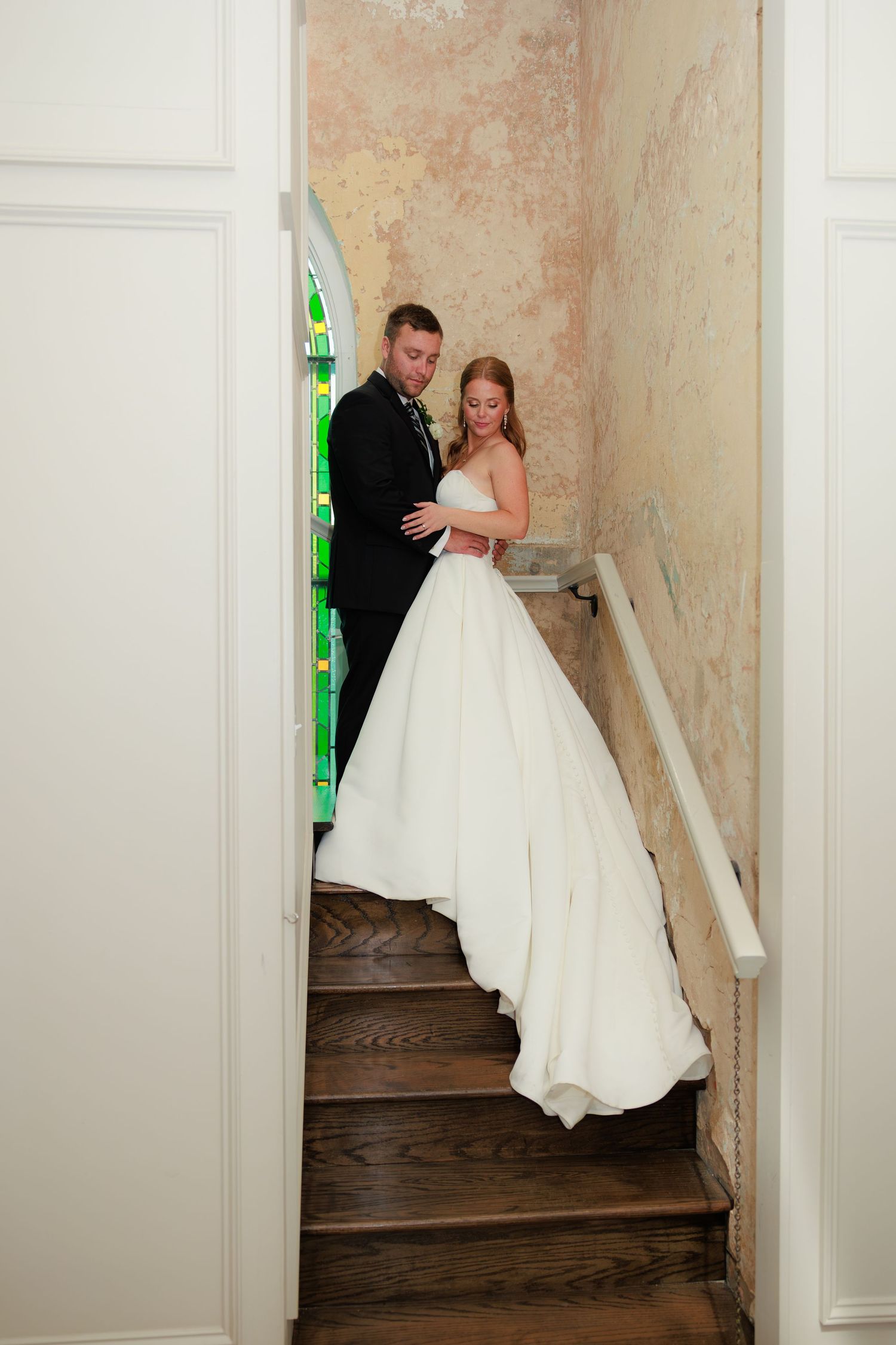 Bride in full white ballgown and groom embrace on wooden staircase near stained glass window indoors.