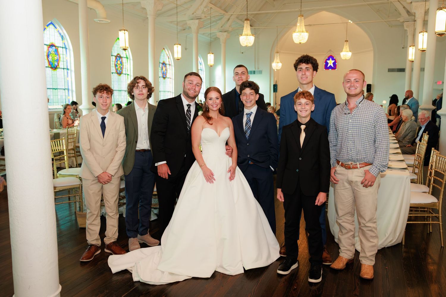 Bride and groom pose with group of young men in elegant white wedding venue with stained glass windows.