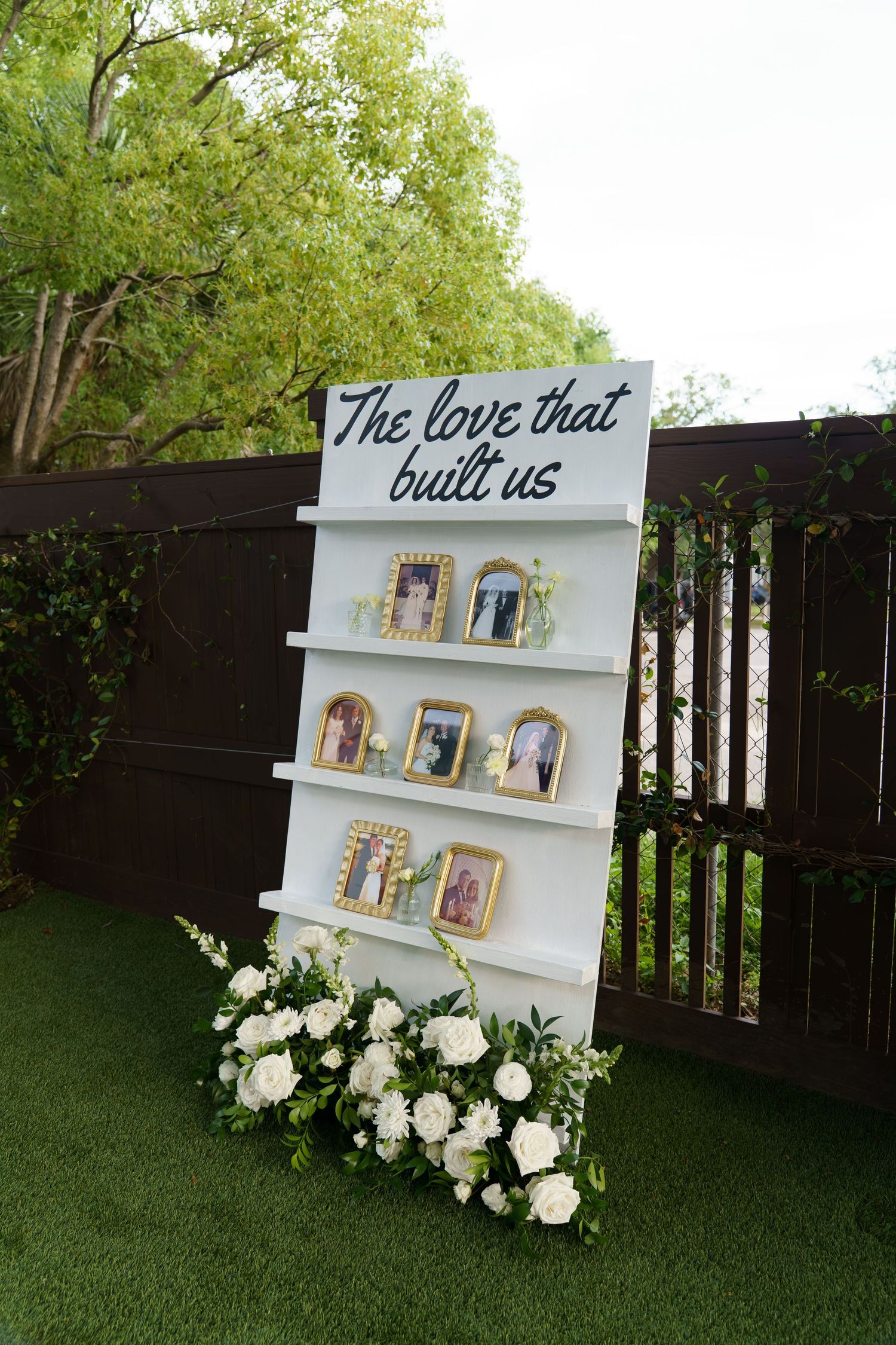 White wedding display shelf with framed photos and sign reading The love that built us, adorned with white floral arrangements.