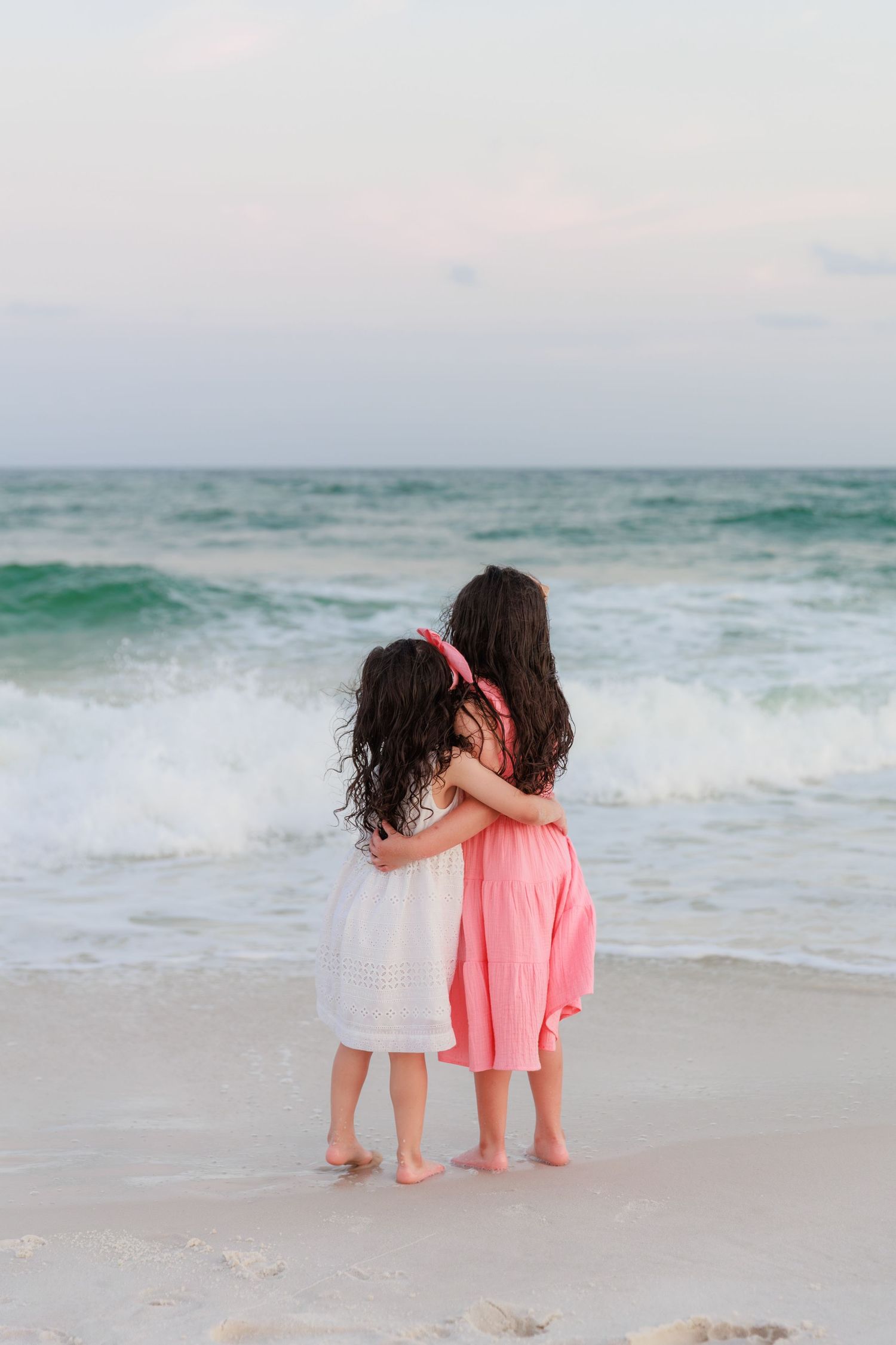 Two young sisters in white and pink dresses hug while watching ocean waves on the beach.