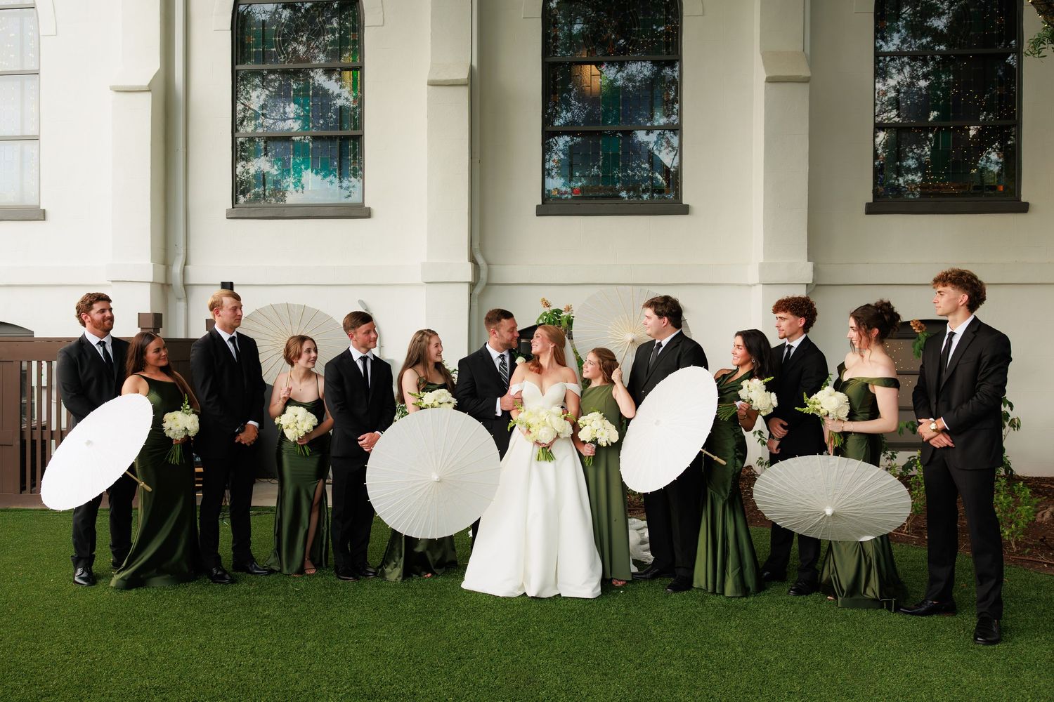 Wedding party in green dresses and black suits holding white parasols outside a white church.