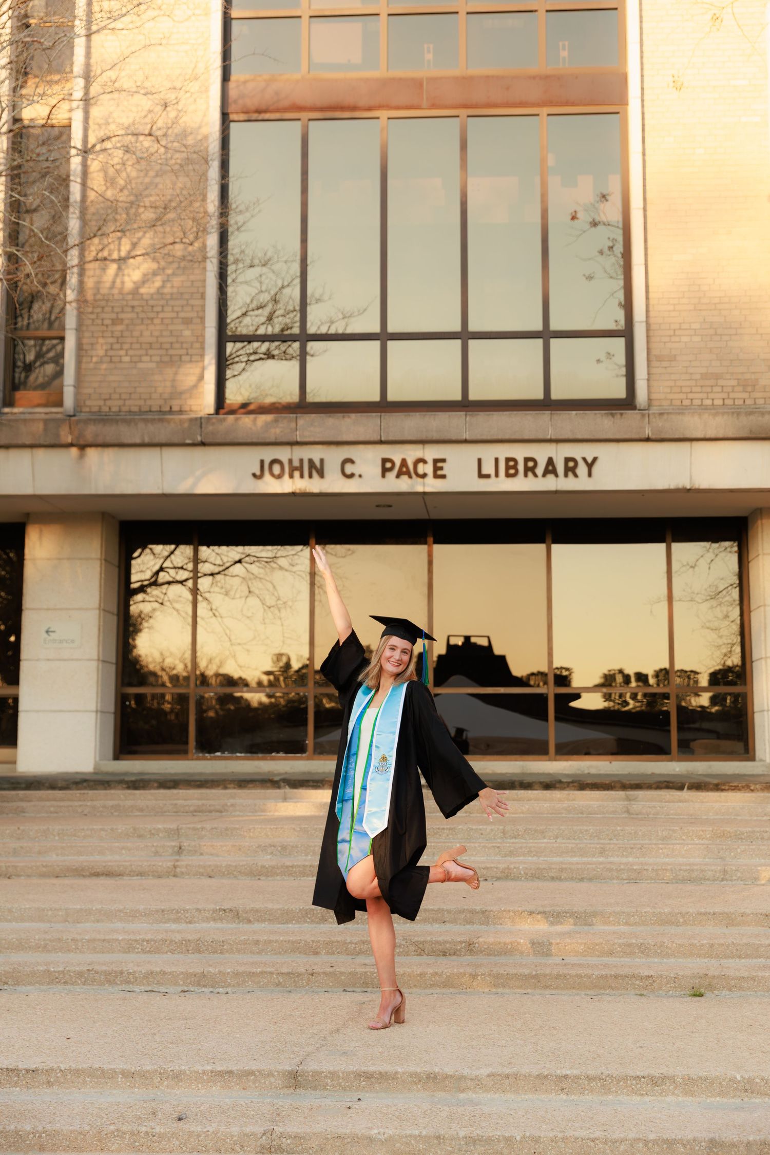 Graduate strikes celebratory pose in academic regalia outside John C. Pace Library on graduation day.