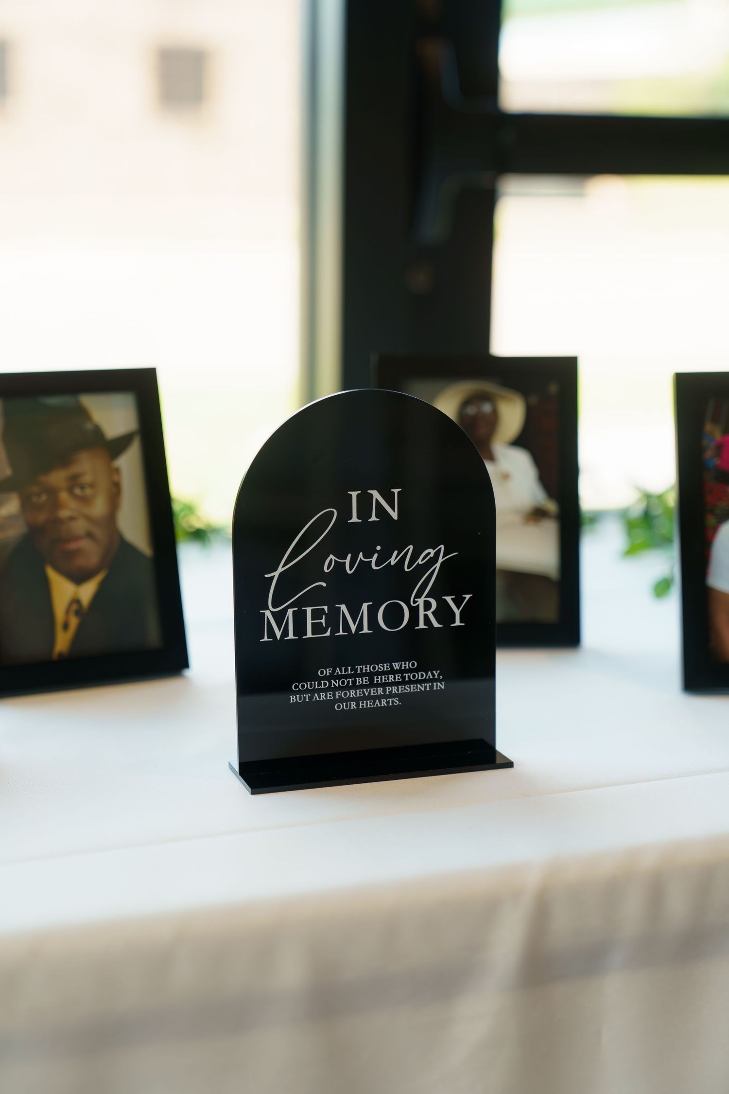 A black 'In Loving Memory' memorial sign stands on a white surface with framed photographs in the background.