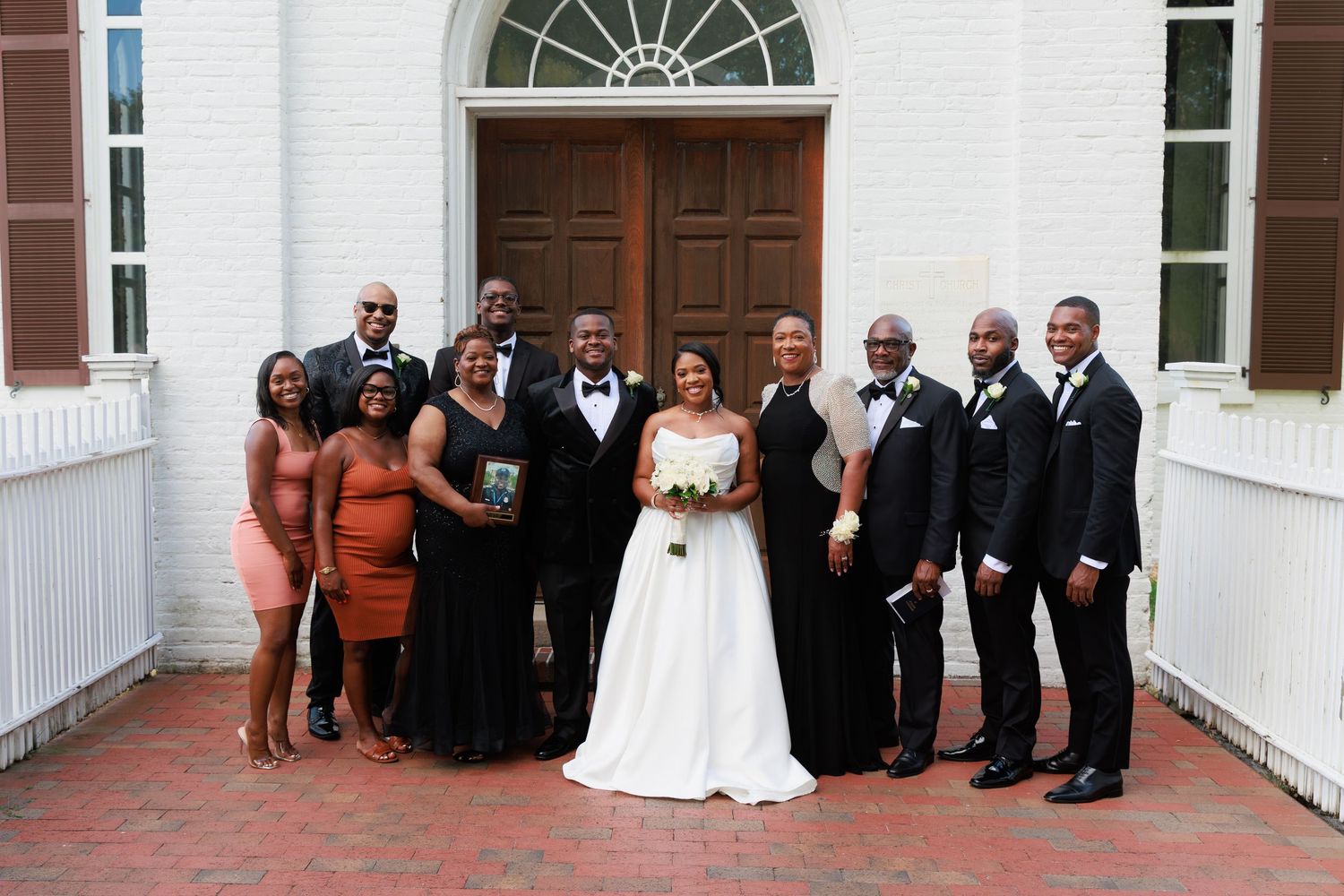 Wedding party in formal attire arranged for group photos outside a white church with arched windows.