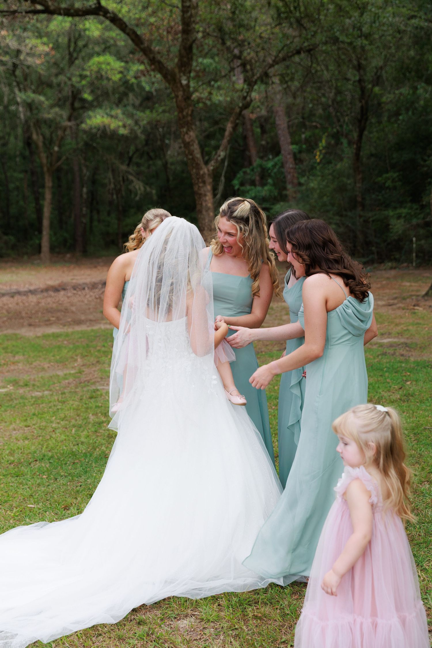 Wedding party members in mint green dresses and a flowing white gown gather for photos in a wooded outdoor setting.