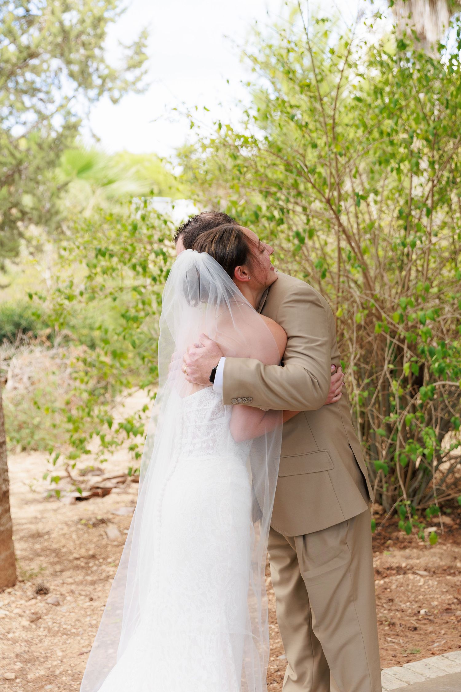 A couple shares a romantic embrace outdoors surrounded by green foliage during a wedding photo session.