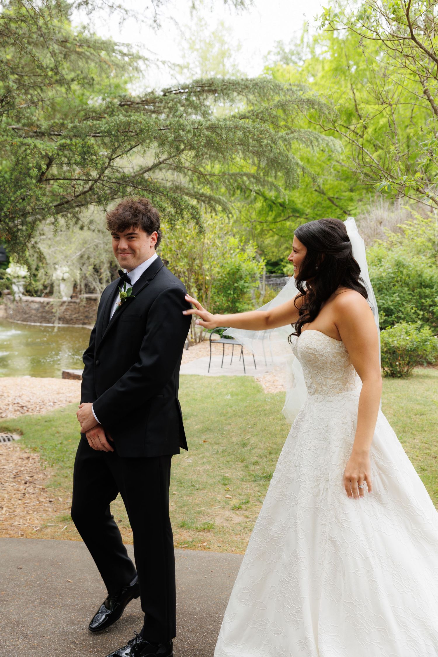 Bride in white gown touches groom's shoulder in black tuxedo during first look in lush garden.