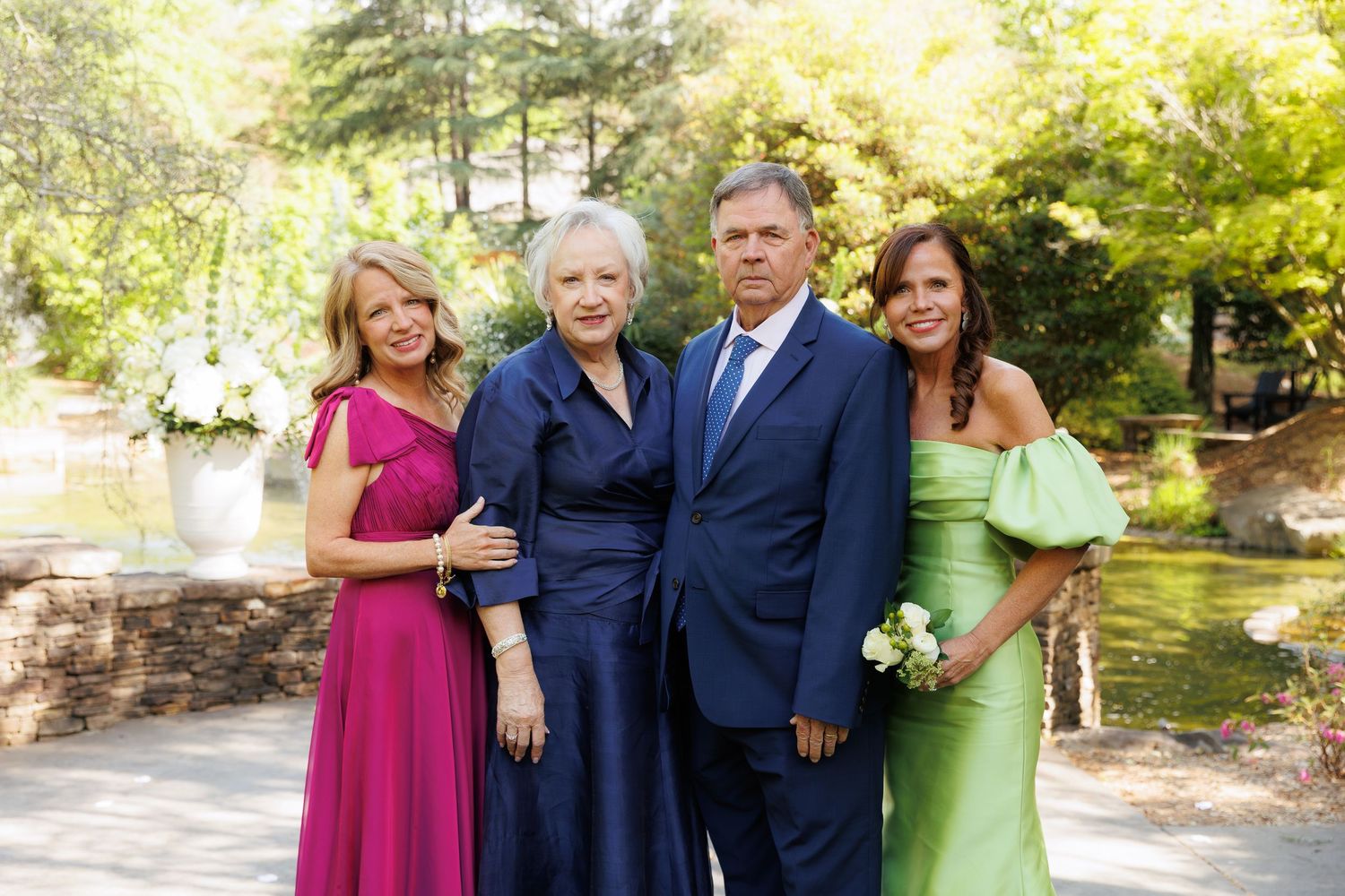 Four elegantly dressed people posing outdoors at a garden event, wearing formal attire in pink, blue, and green.