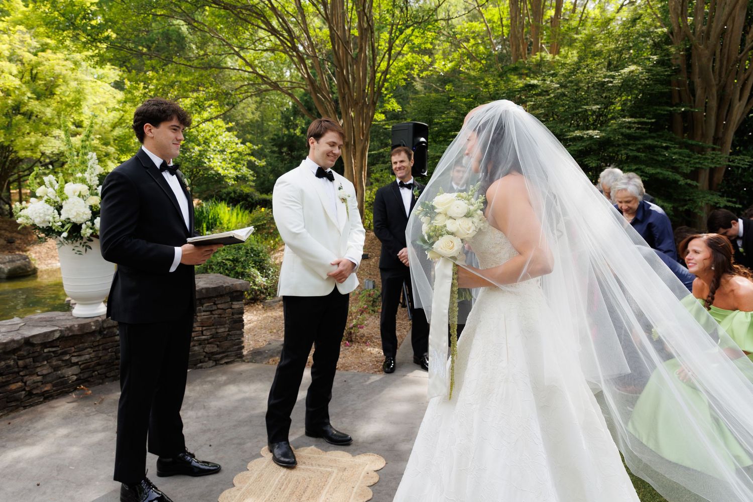 Outdoor wedding ceremony with bride in white gown and veil holding bouquet, groom in white jacket, officiant in tuxedo.