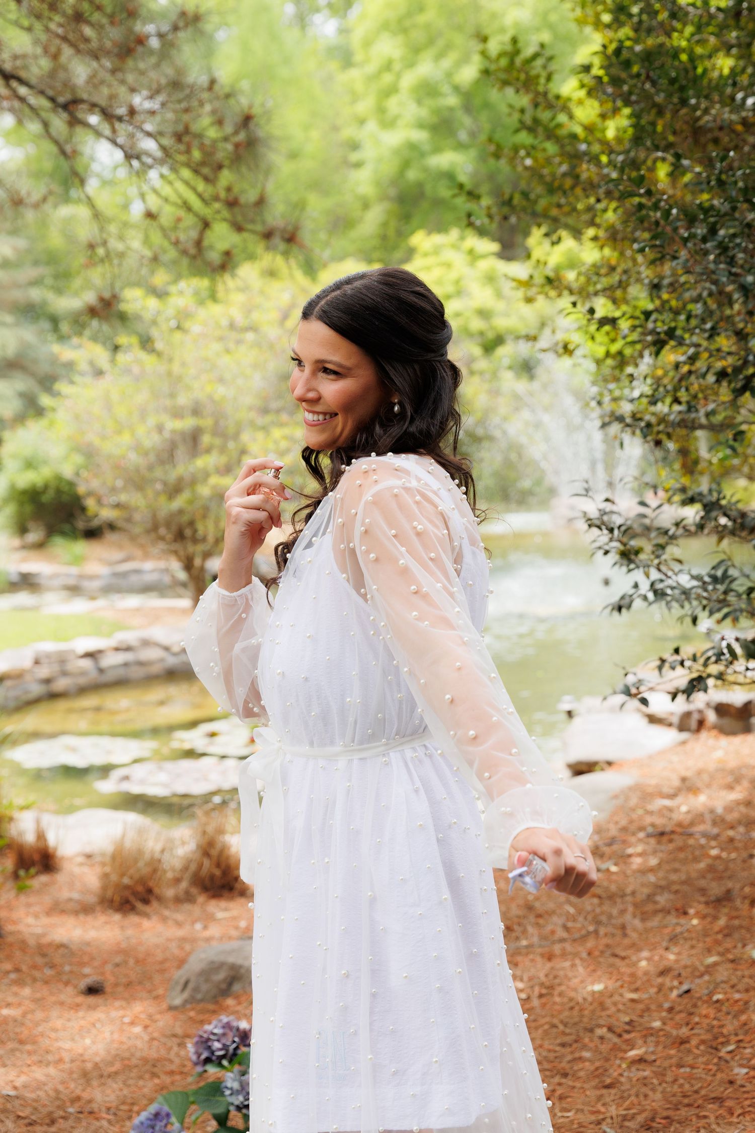 Smiling bride in white lace long-sleeve dress poses in a lush garden with a pond and waterfall backdrop.