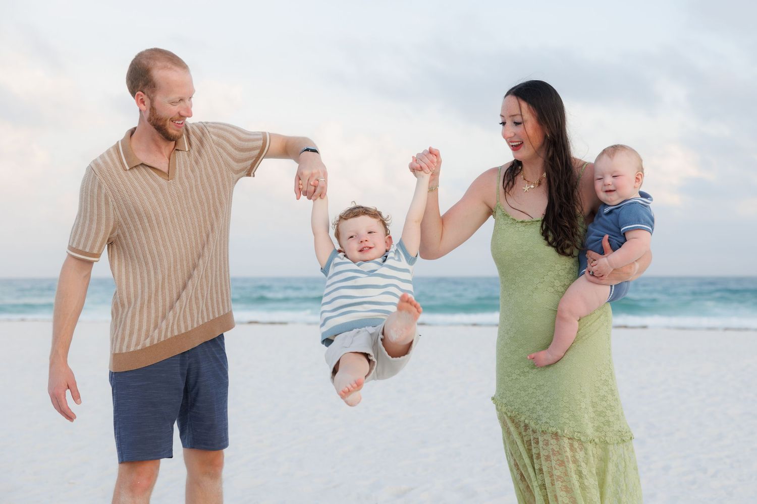Joyful parents swing toddler on beach while mom holds baby, ocean backdrop at dusk.