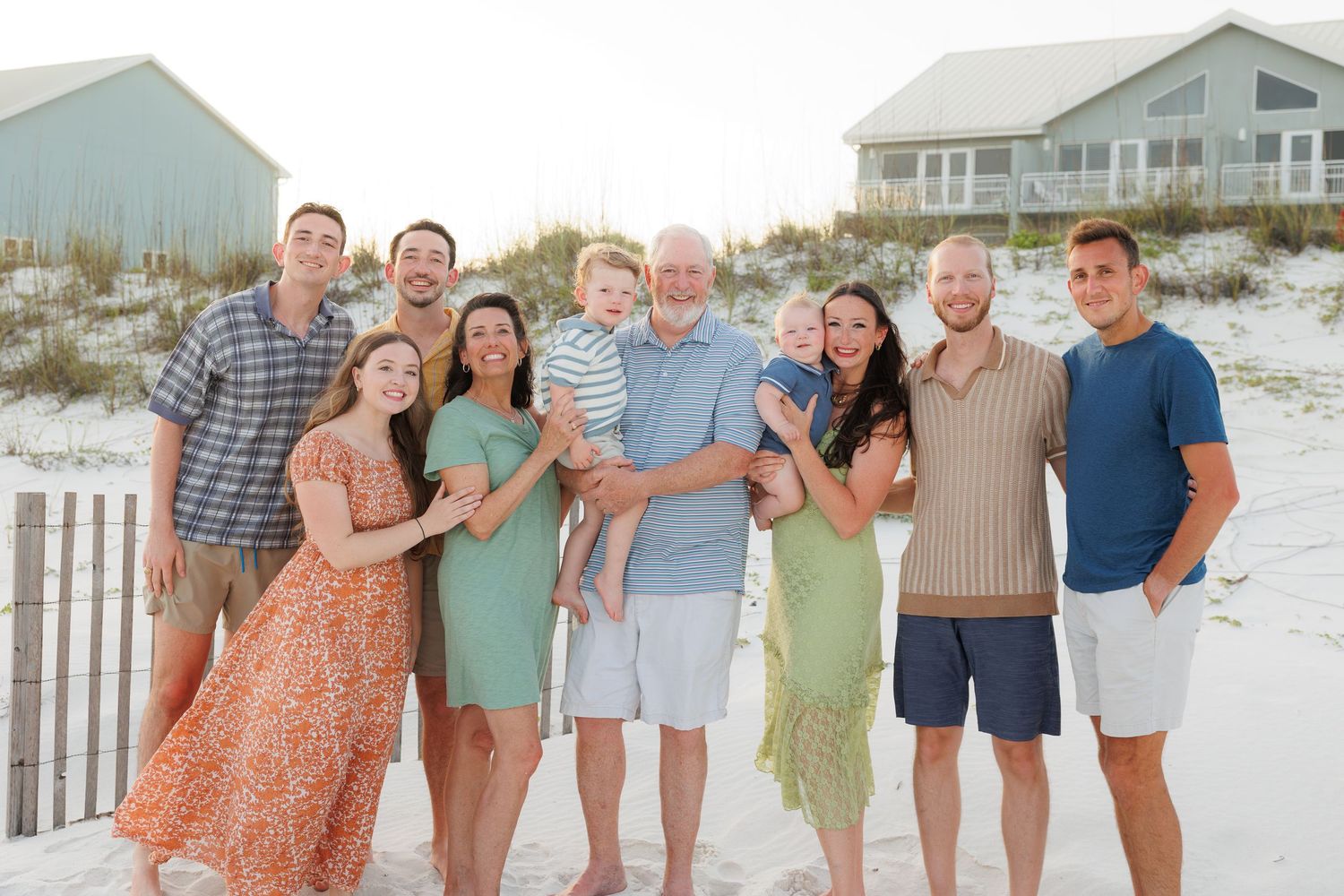 Multi-generational family smiling together on a sandy beach with dunes and coastal homes in the background.
