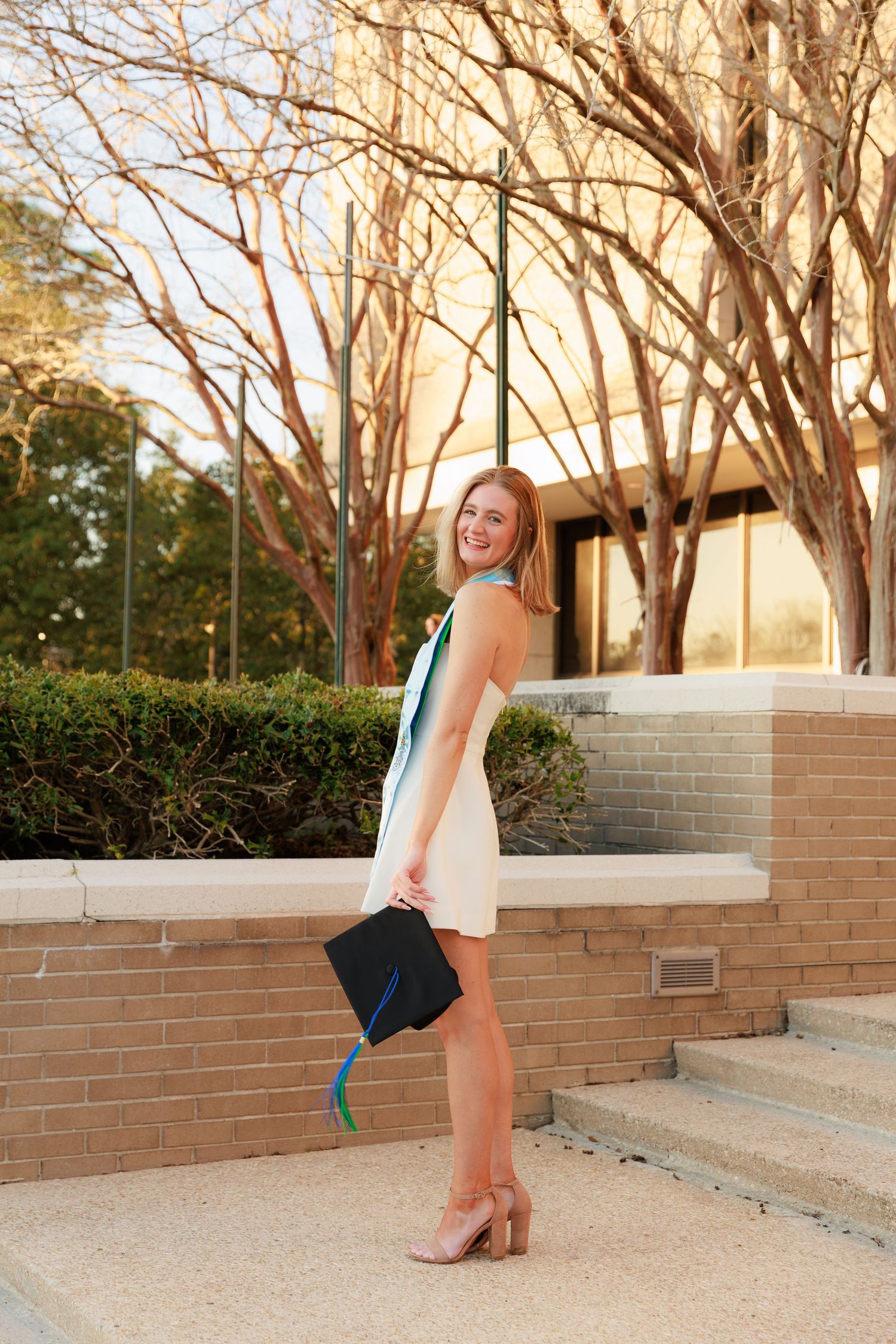 A person in a white sleeveless dress and black clutch poses against a brick wall during golden hour lighting.