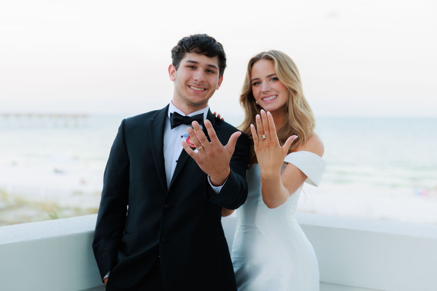 A couple in wedding attire showing their rings on a balcony with ocean view in the background.