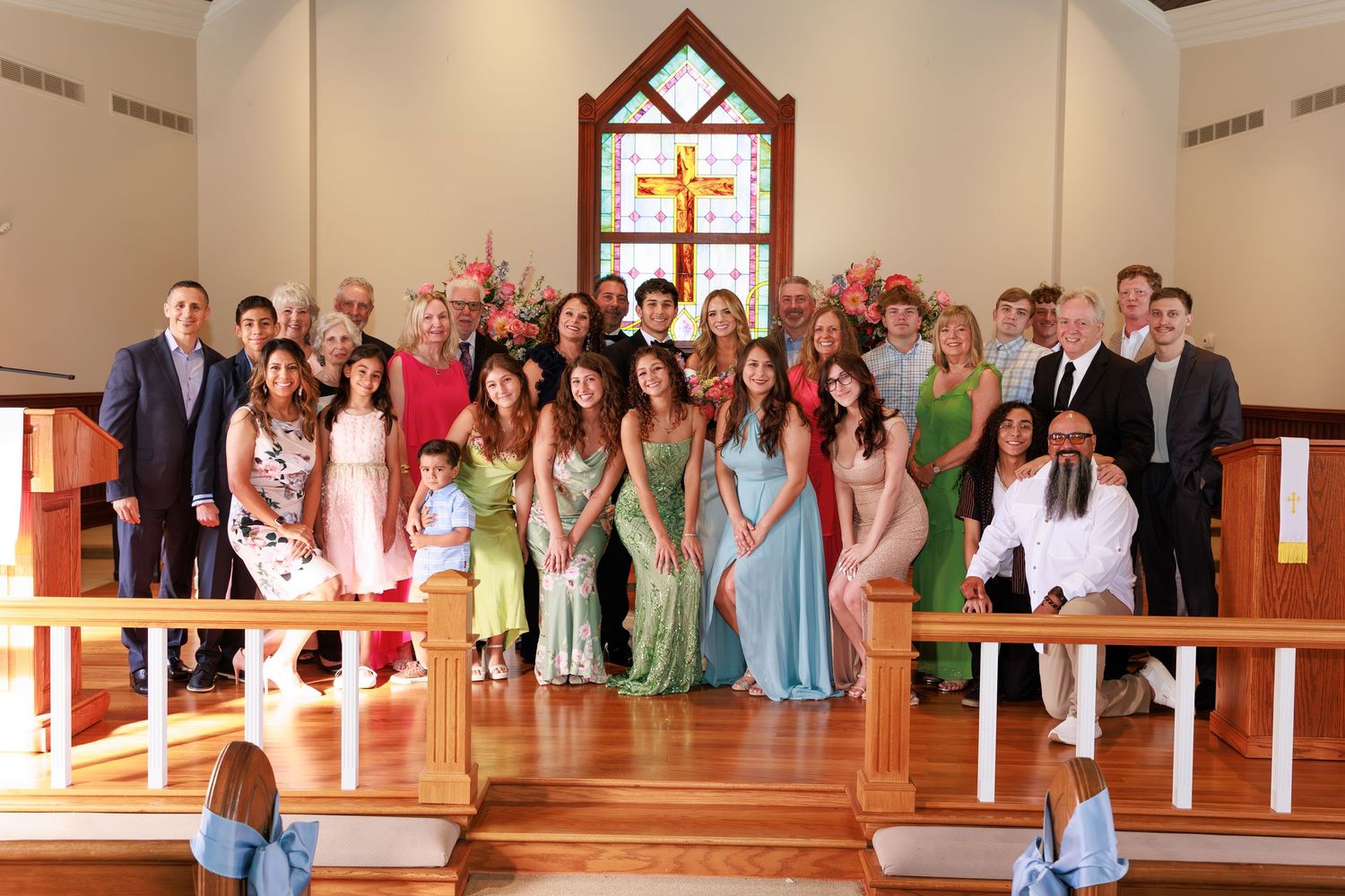 Group of wedding guests pose for formal portraits in a church sanctuary with wooden pews and a stained glass window.