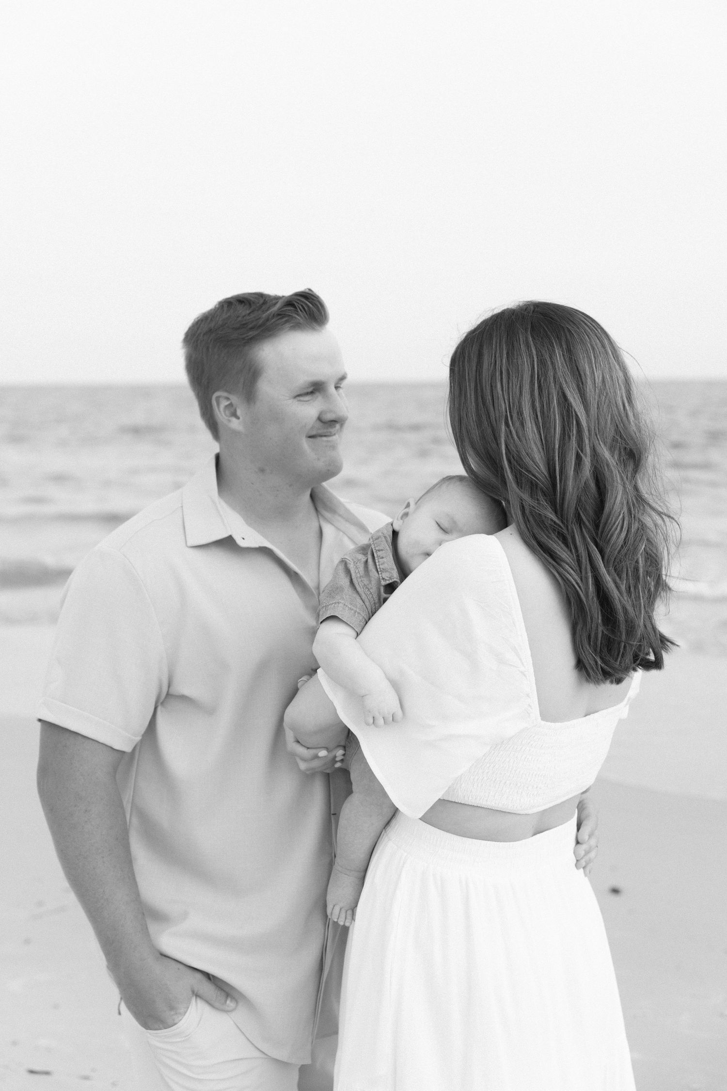 Black and white beach photo of a couple holding their newborn baby by the ocean.