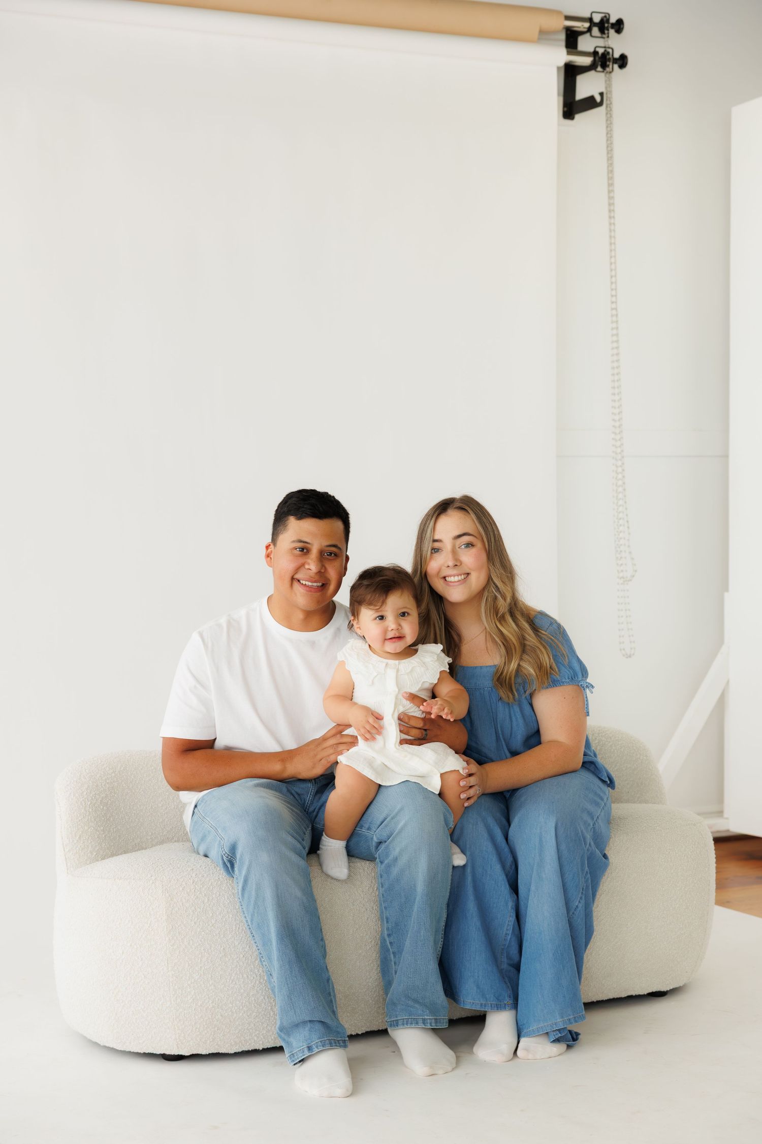A happy family of three poses on a white sofa, parents in denim outfits holding their baby girl in a bright studio.