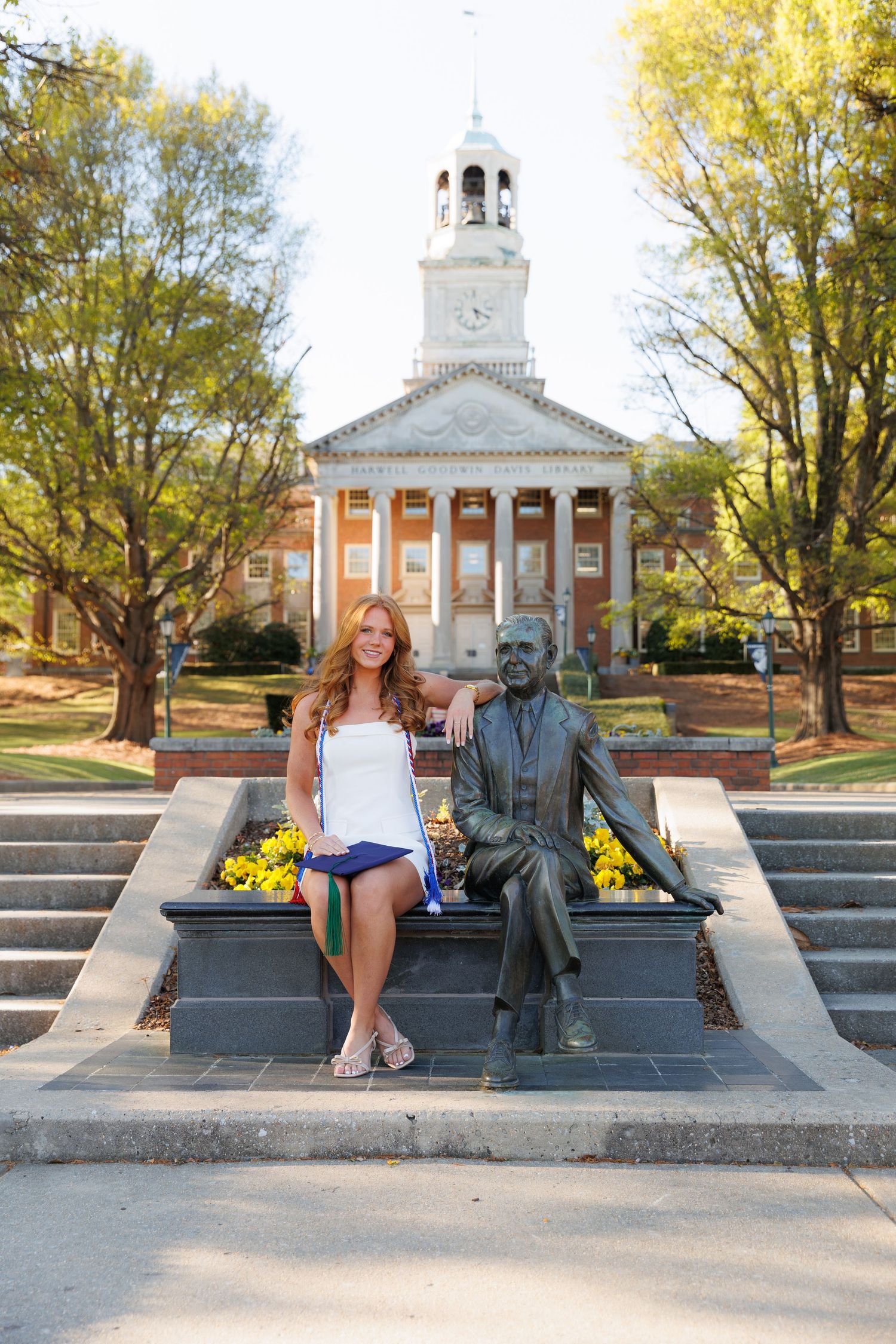 Young woman poses beside a bronze statue on campus steps with a historic brick building and clock tower in the background.