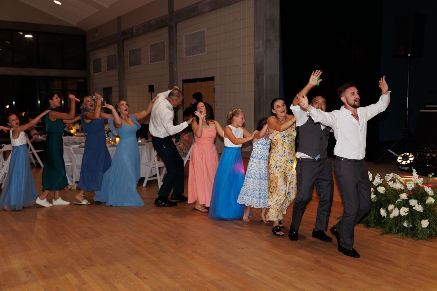 Wedding guests perform a line dance in formation with bridesmaids in blue dresses on a dance floor.
