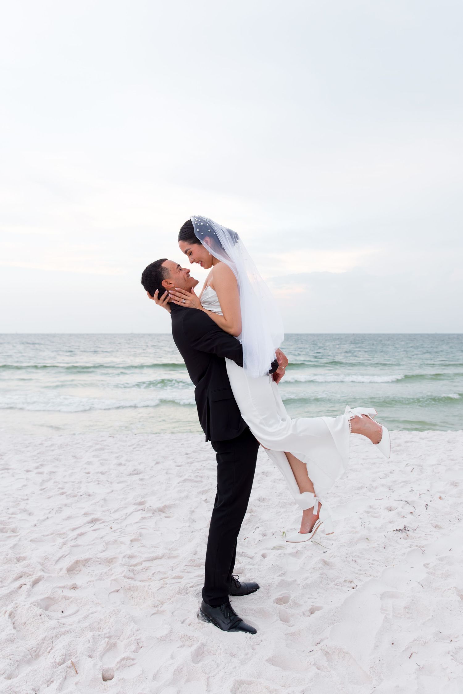 Couple in wedding attire embrace playfully on white sandy beach.