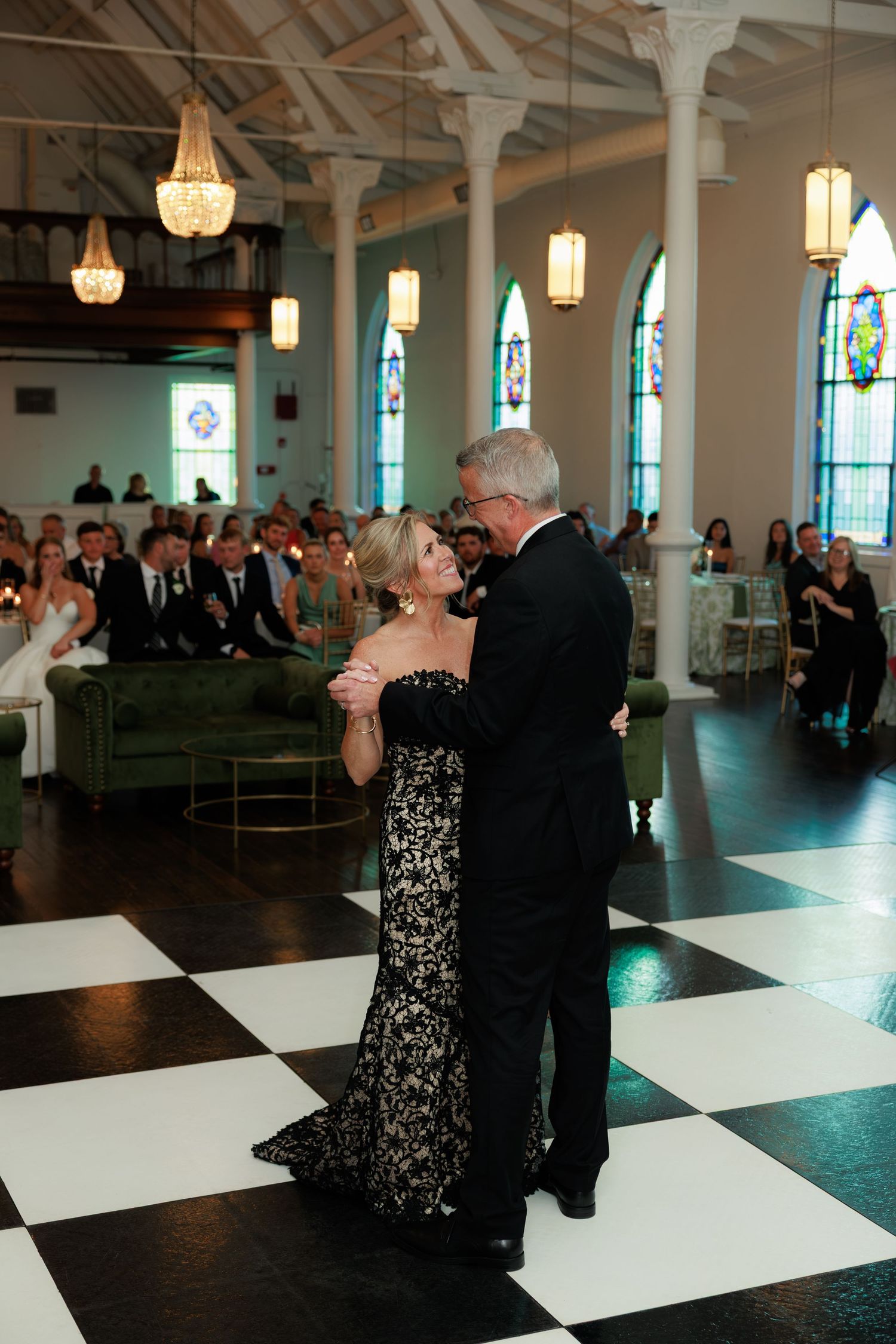 Elegant couple dancing on black and white checkered floor at wedding reception in church venue with stained glass windows.