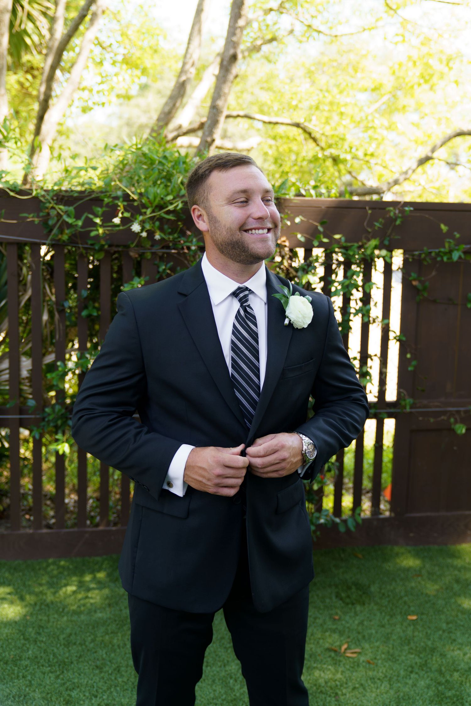 Smiling groom in dark suit with striped tie and white boutonniere buttoning jacket outdoors.
