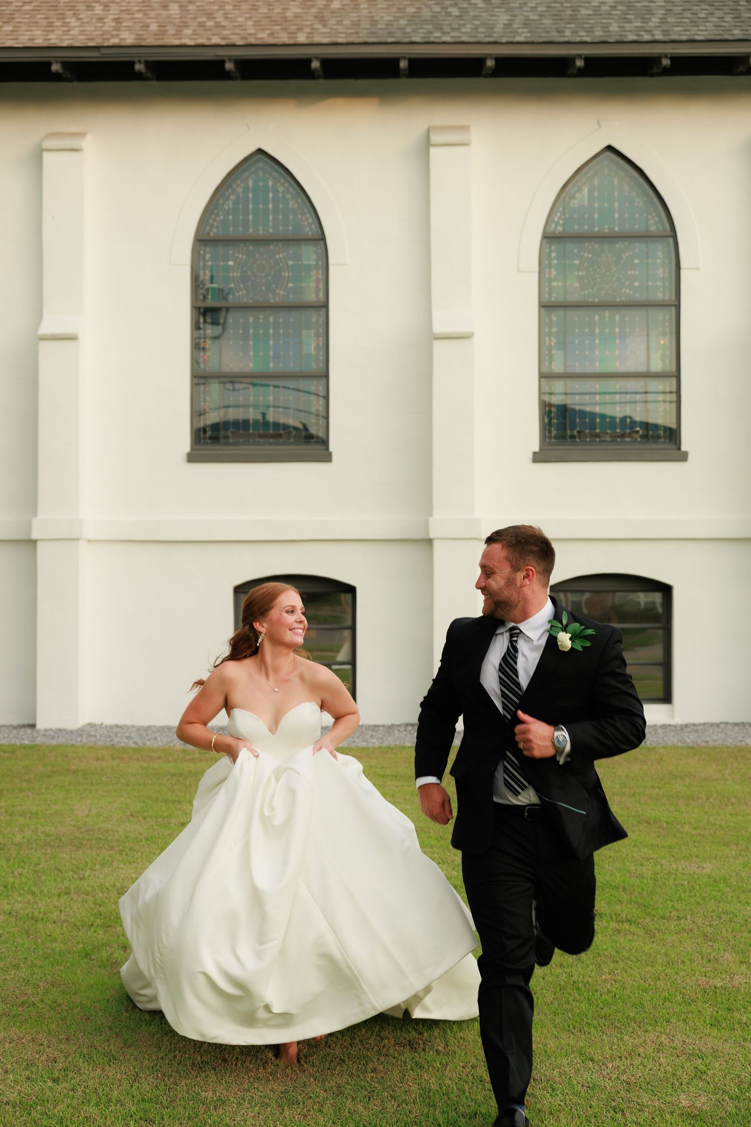 Joyful bride and groom run and laugh together on lawn outside white Gothic church with arched windows.