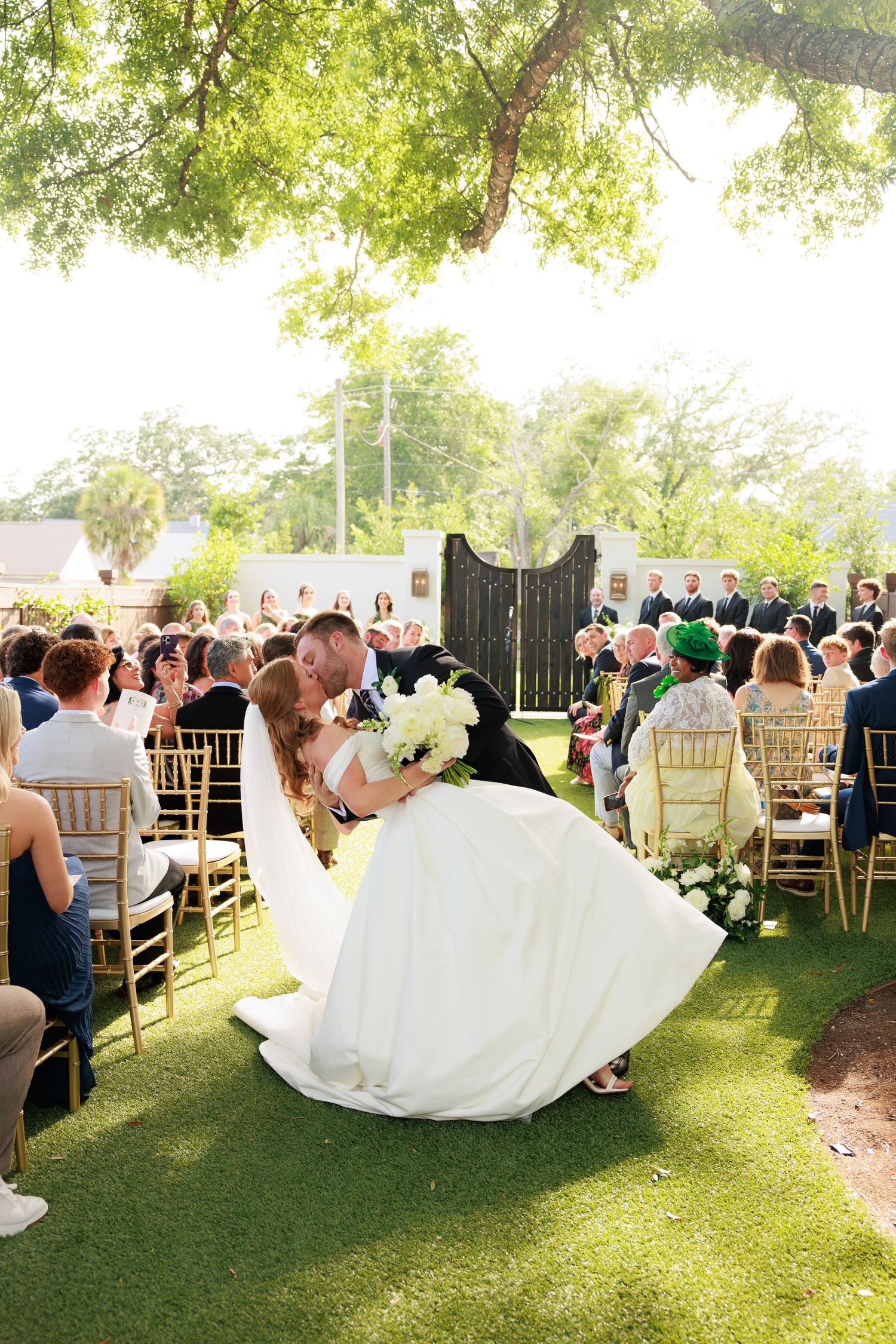 Groom dips bride for a kiss mid-aisle at sunny outdoor wedding.