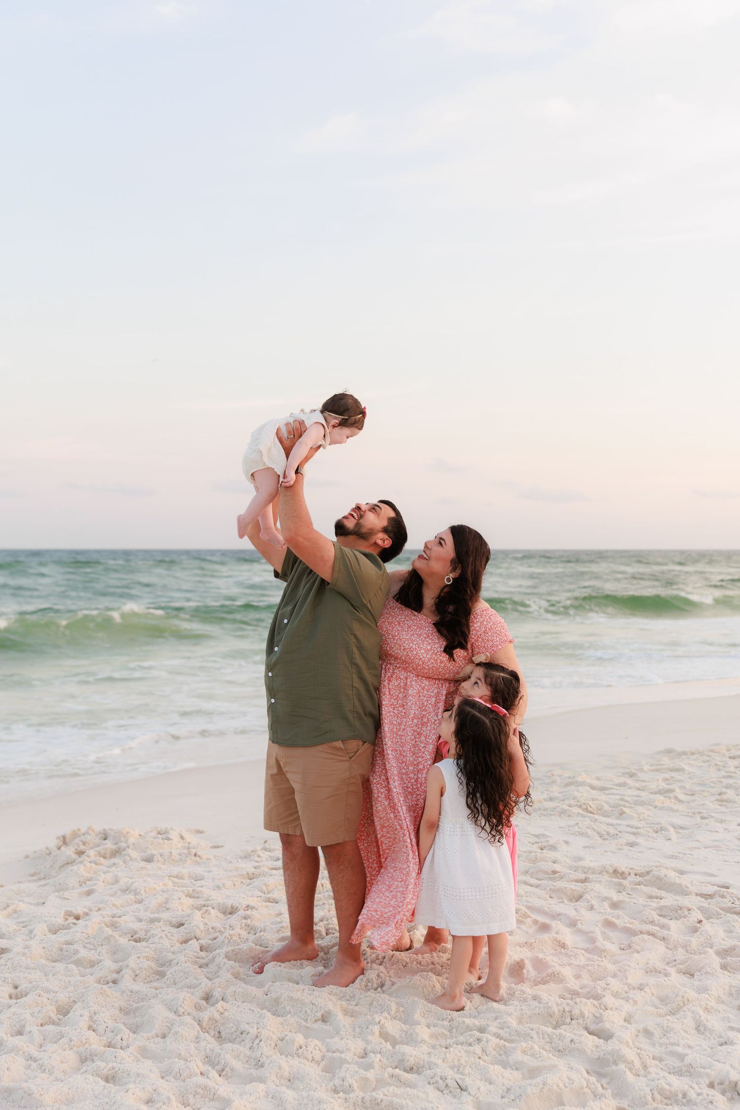 Dad lifts baby up while mom and daughter hug together as a family on a beach at sunset.