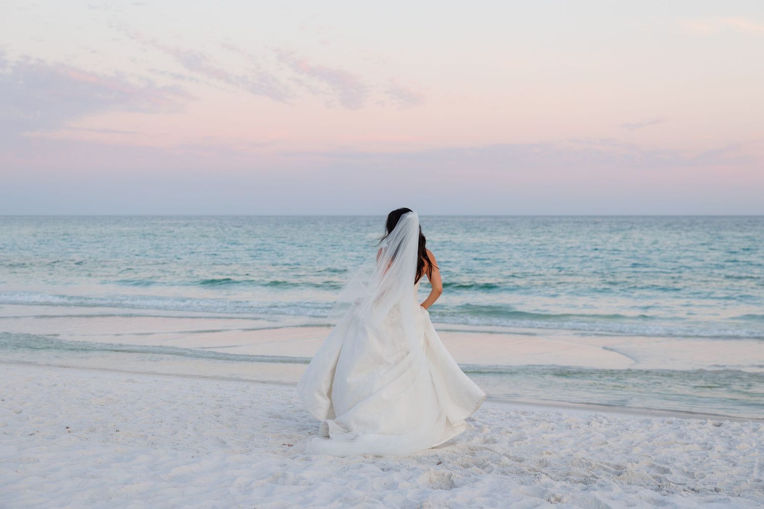 Bride in white wedding gown and veil stands on sandy beach at sunset with calm turquoise ocean waves.