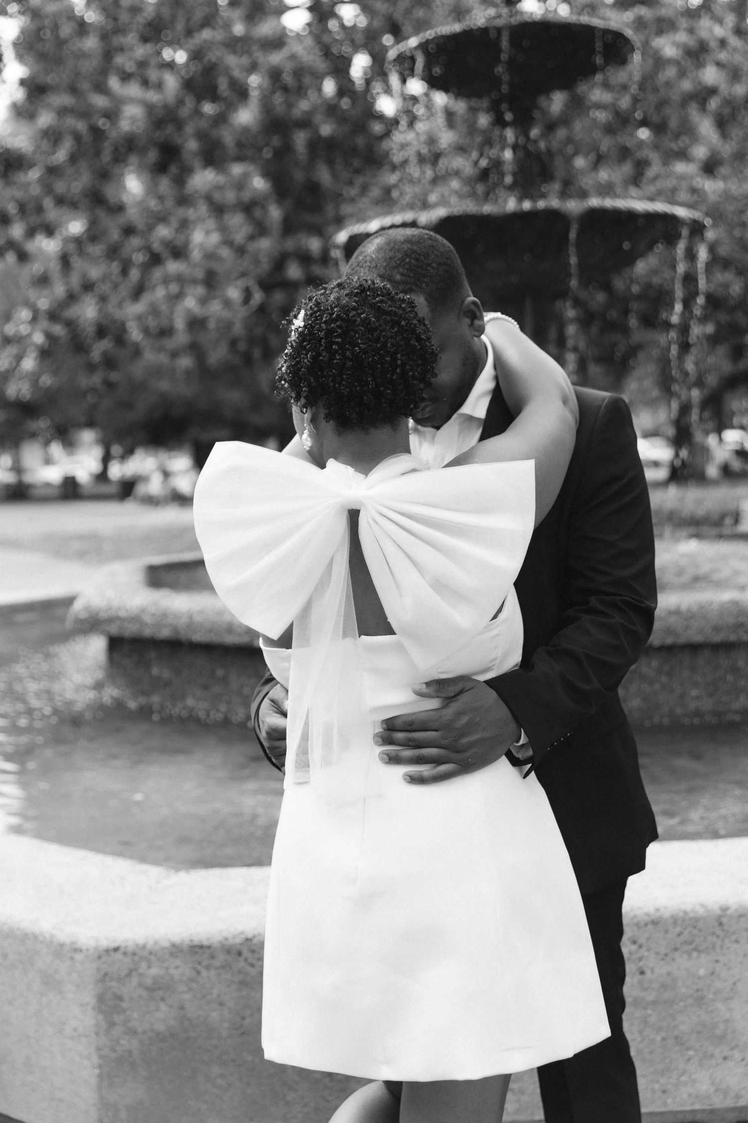 Black and white photo of a couple embracing and kissing near a fountain, bride wearing a white dress with large bow.