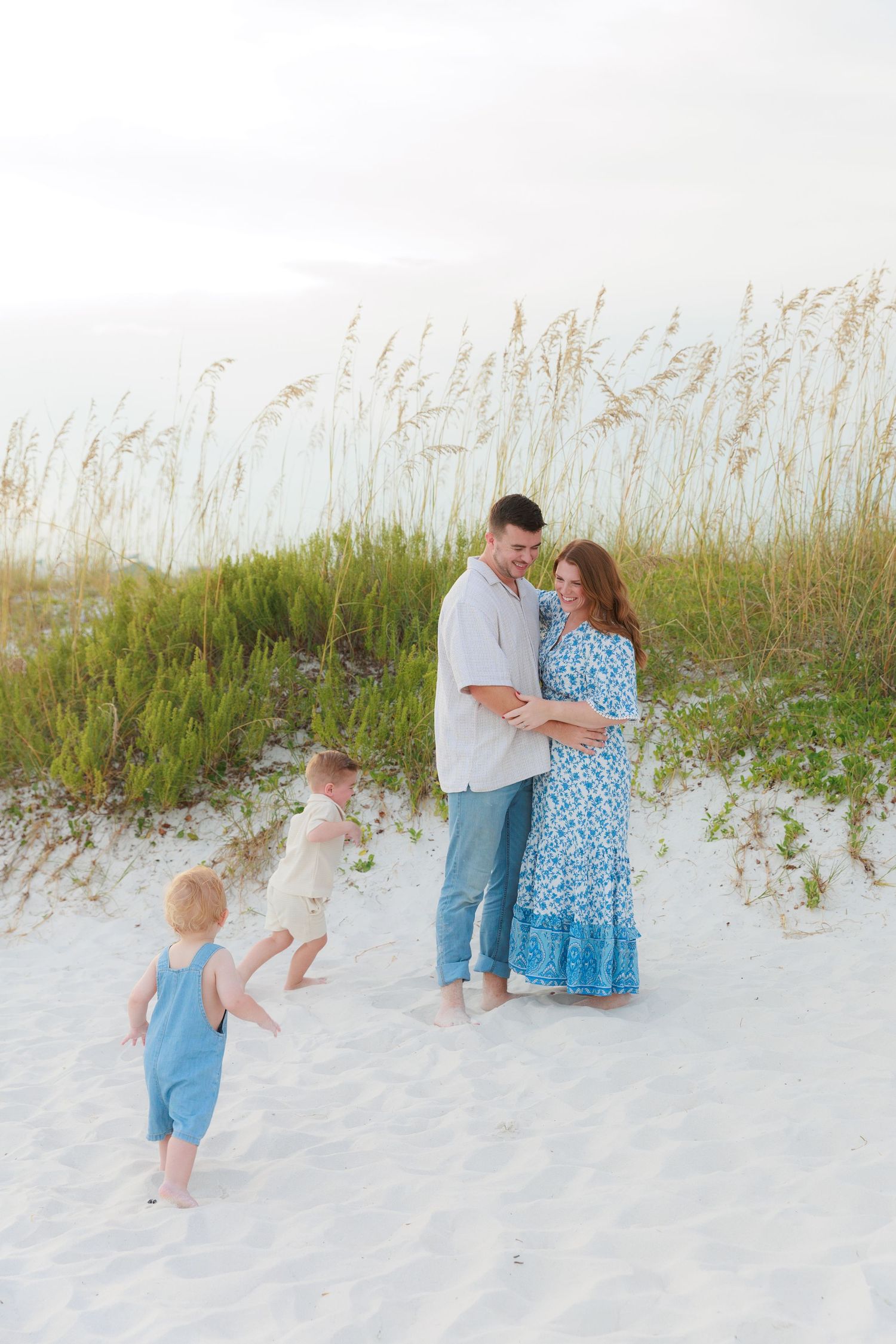 A happy family poses on white sand beach dunes with sea oats, two toddlers running toward smiling parents.