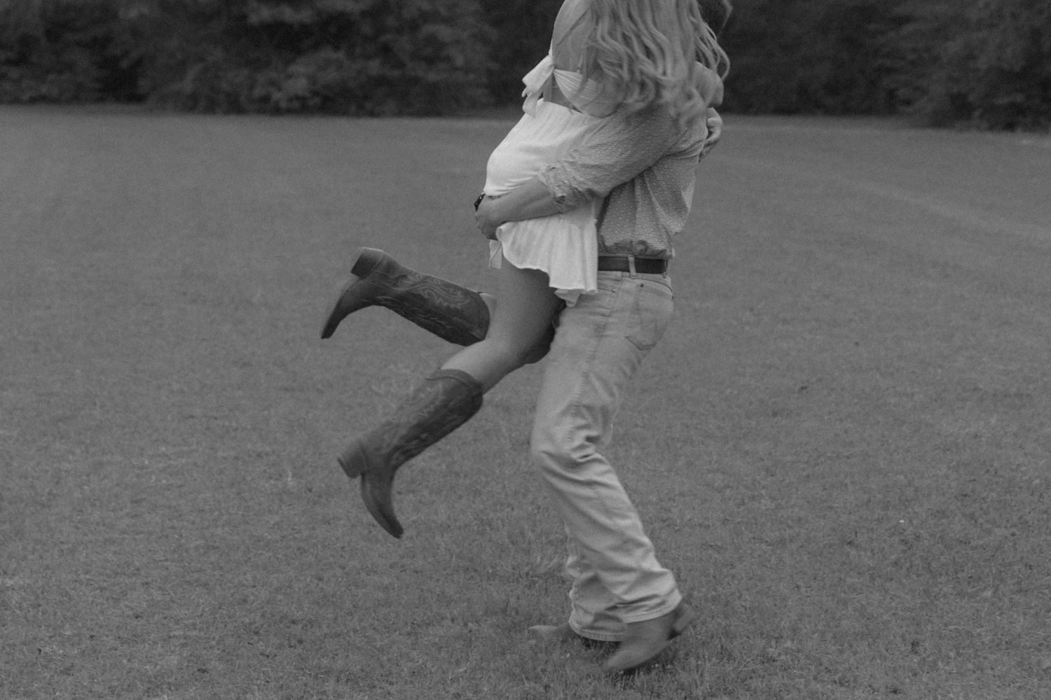 Black and white photo of a couple embracing in a field, woman lifted wearing cowboy boots and a skirt.