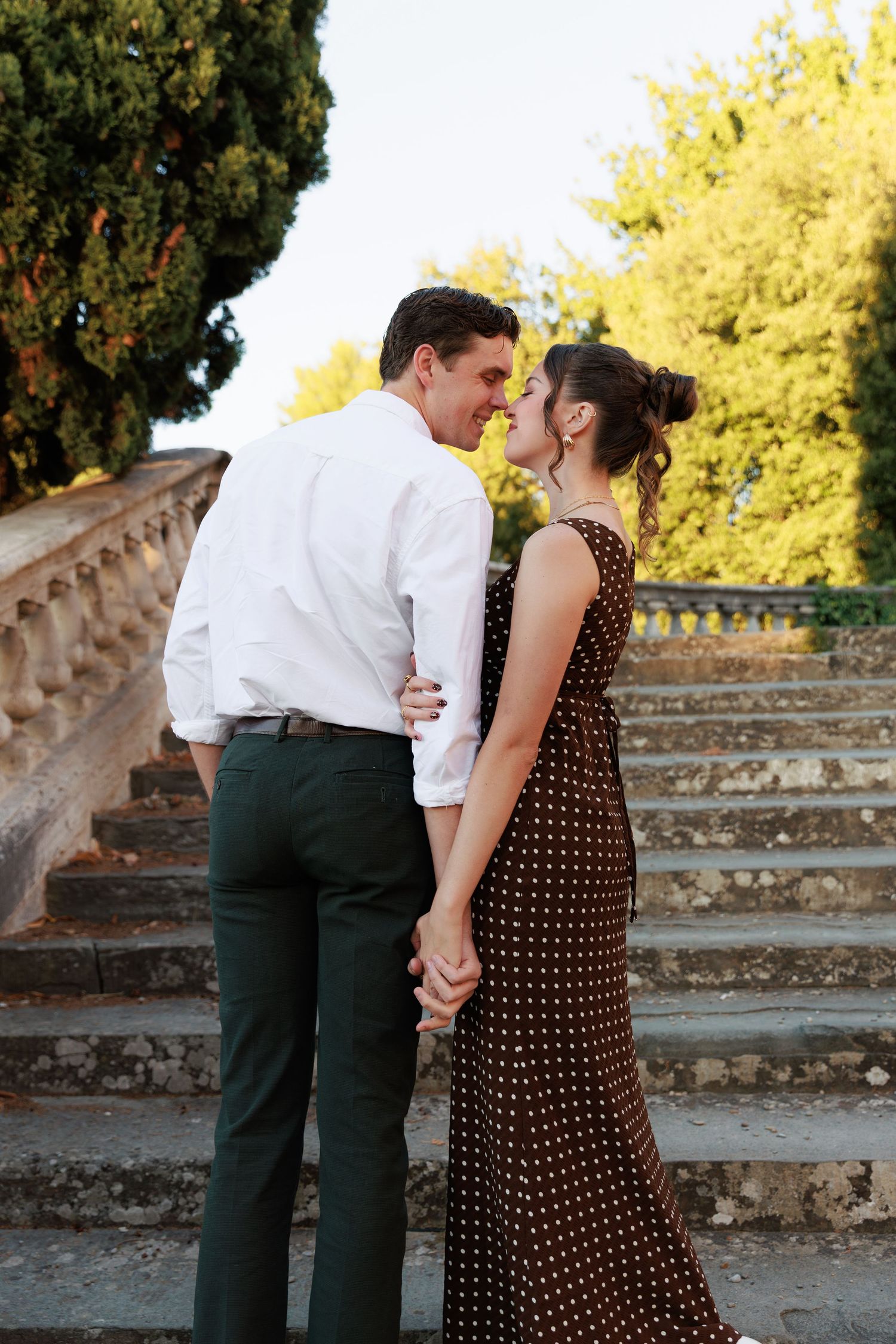 Romantic couple sharing an intimate moment on stone steps, woman in brown polka dot dress, man in white shirt.