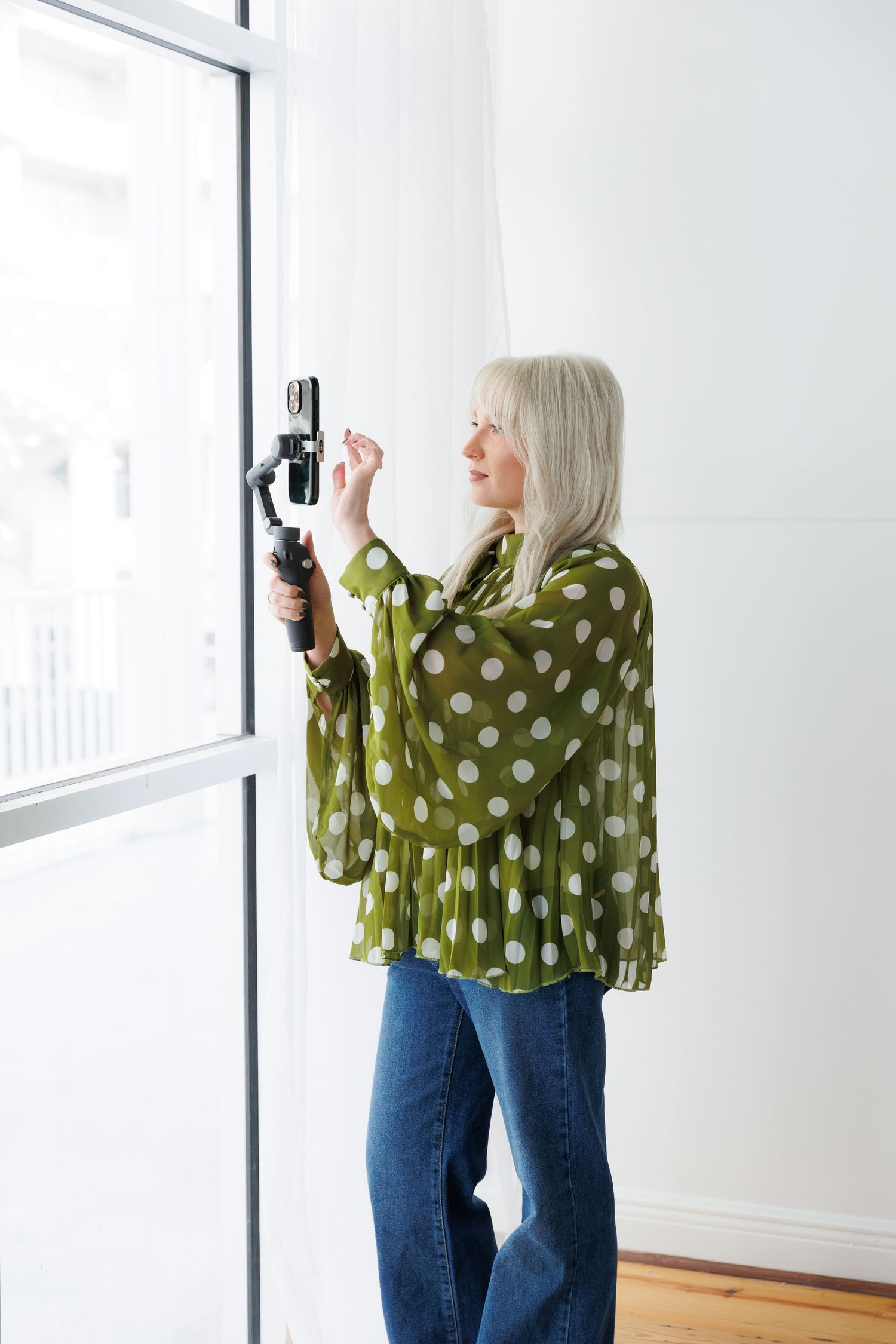 Person wearing green polka dot top and jeans takes photos in bright white interior space.