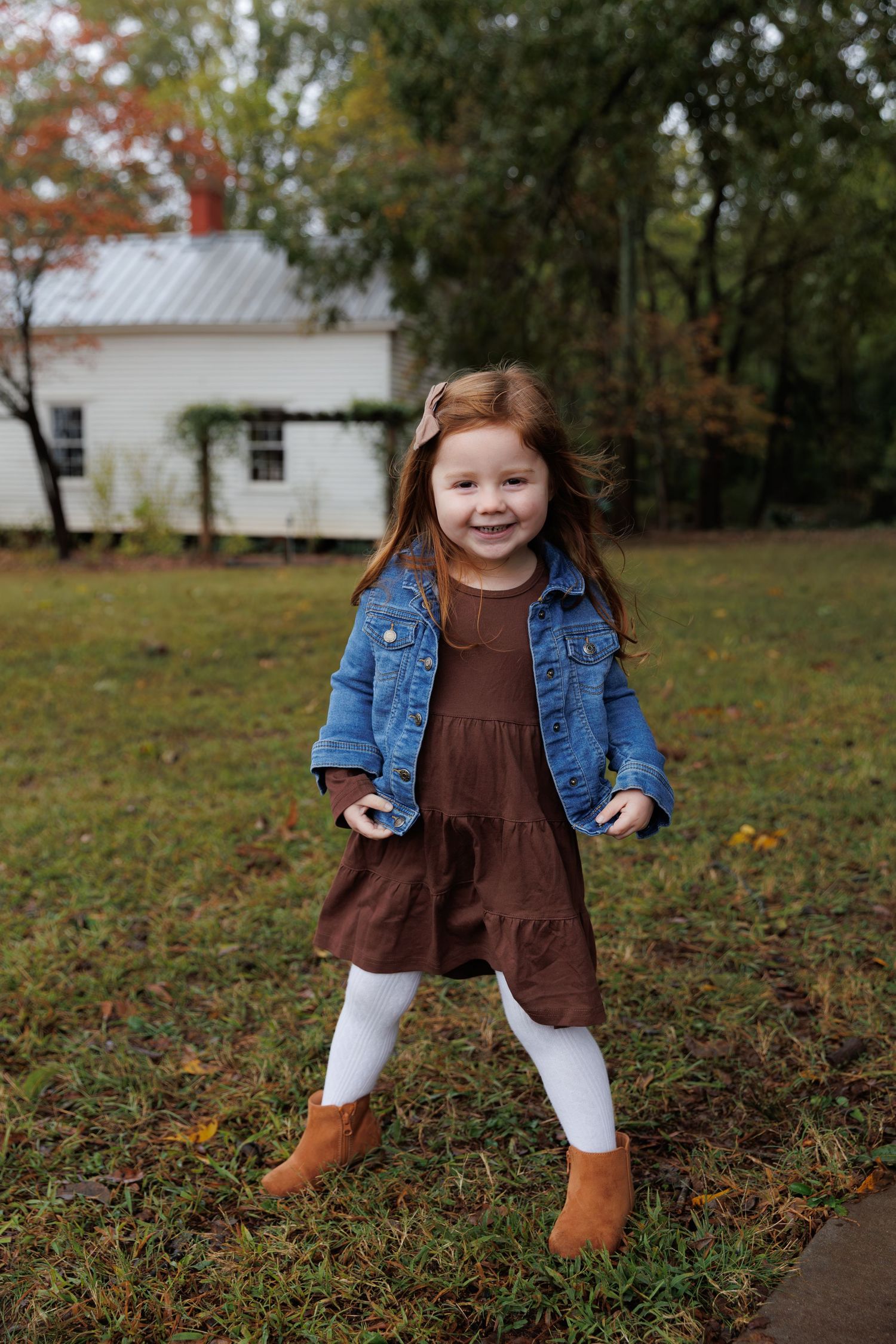 A toddler in a denim jacket and brown dress plays in a yard with autumn leaves and white buildings in background.