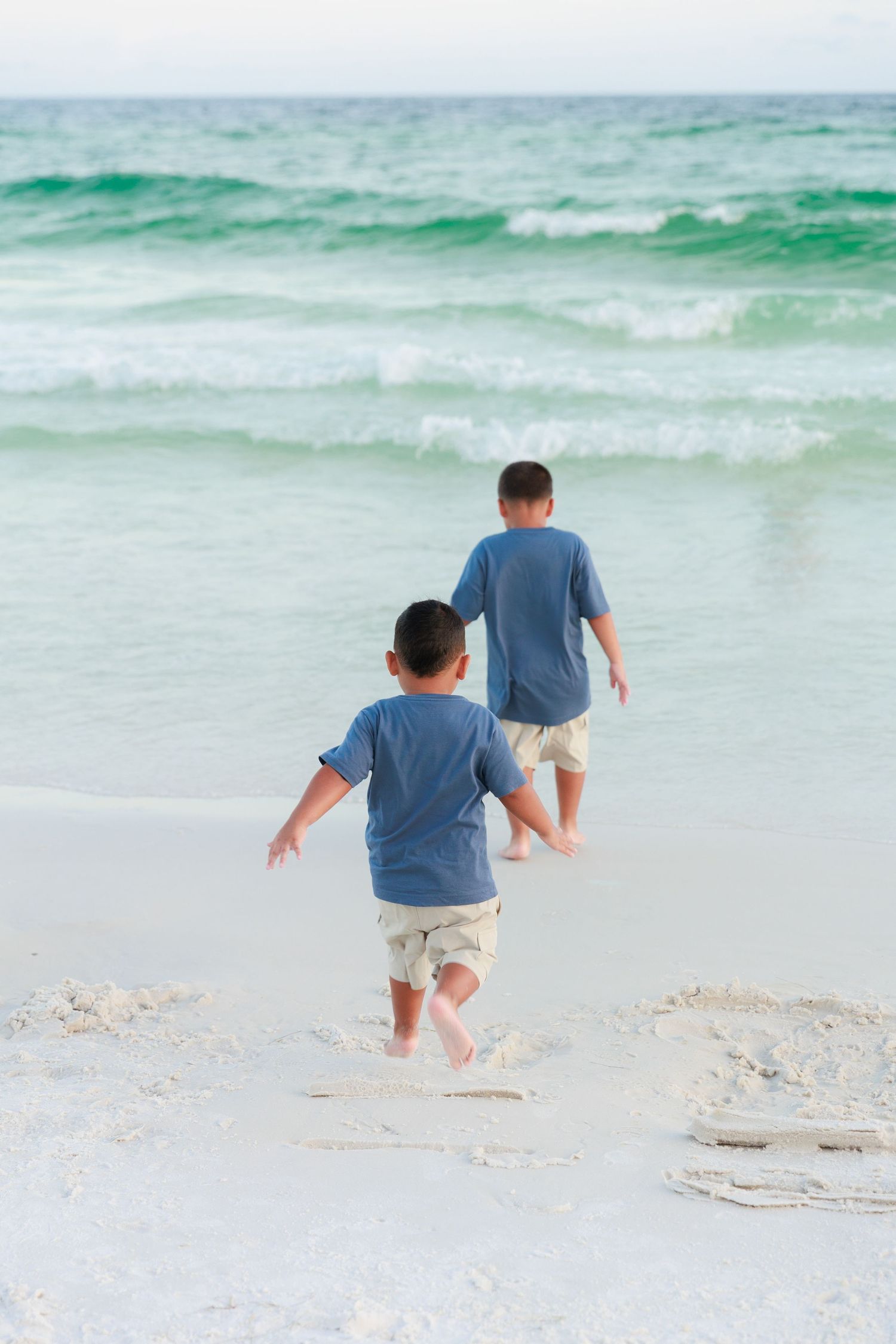 Two people walk along the shoreline as turquoise waves crash on a white sandy beach on a sunny day.