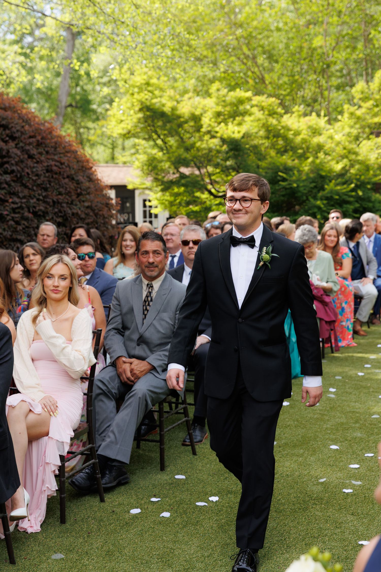 Groom in black tuxedo walks down outdoor wedding aisle lined with seated guests on a sunny day.