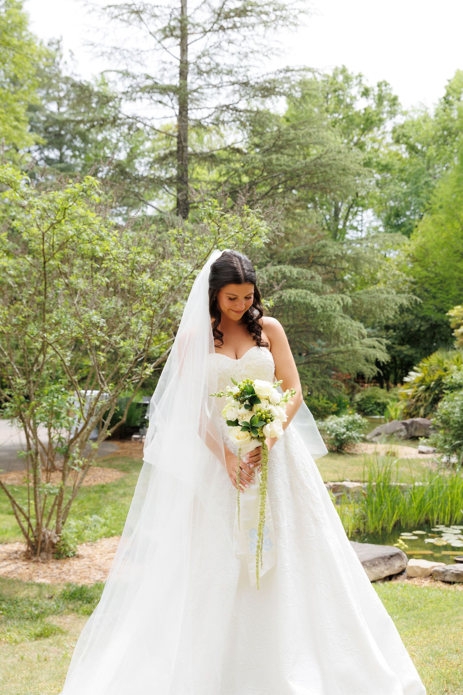 Bride in white strapless gown and veil holds bouquet of white flowers in a lush garden setting.