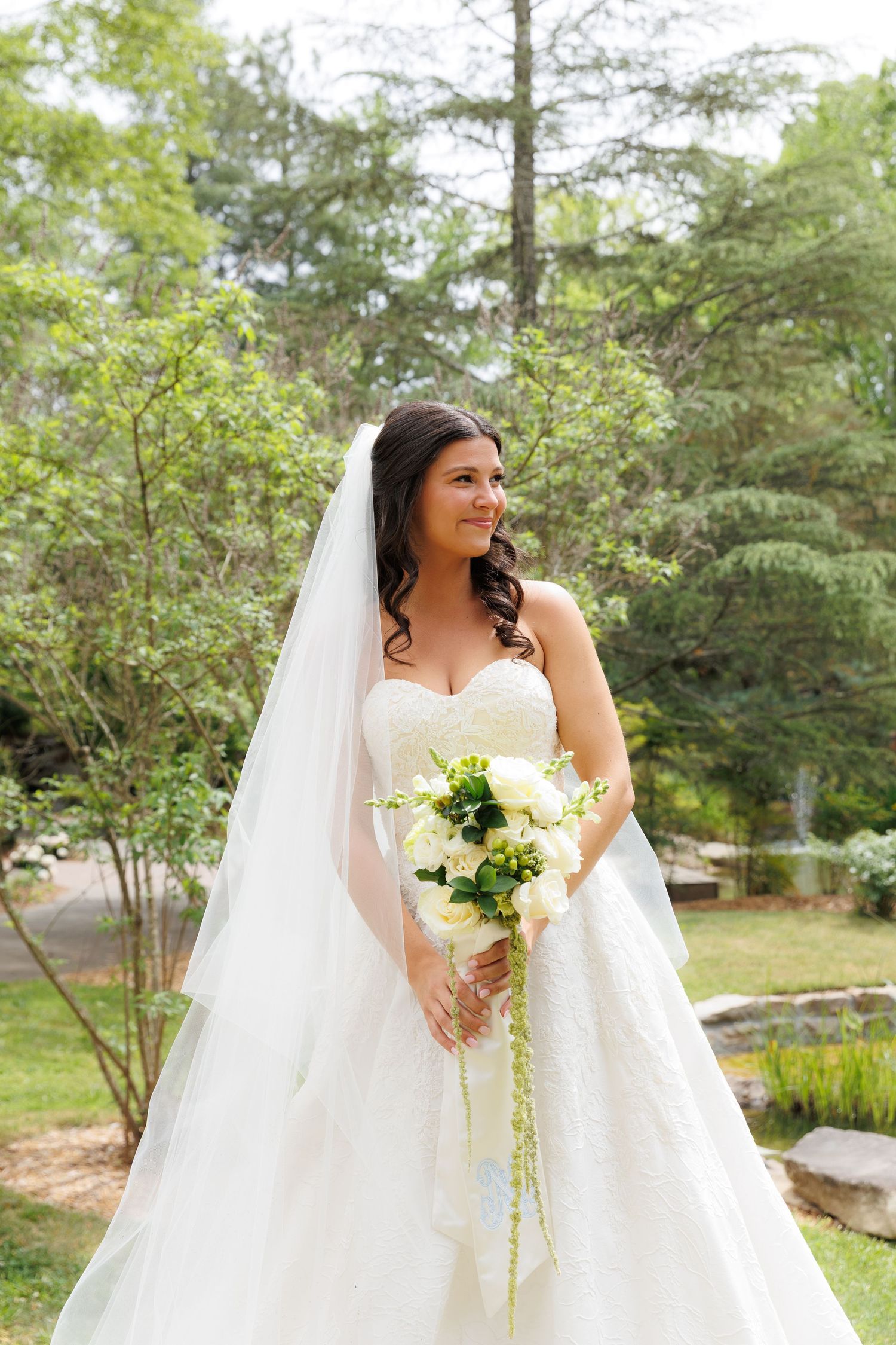 Bride in strapless white ball gown and veil holding white bouquet outdoors in lush green garden.