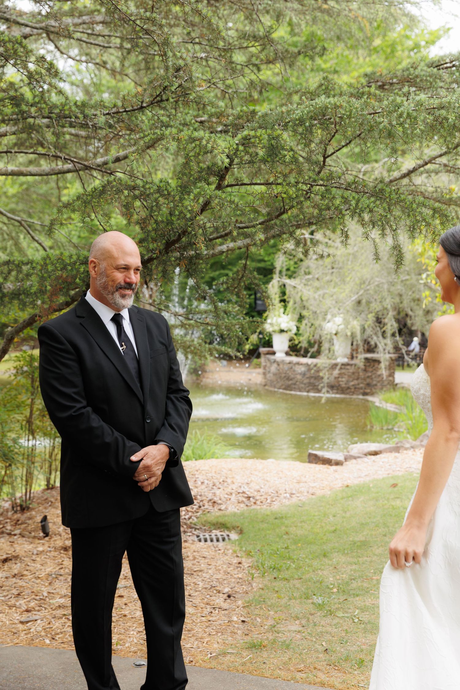 Bald groom in black suit smiles warmly at bride during outdoor wedding first look by garden pond.