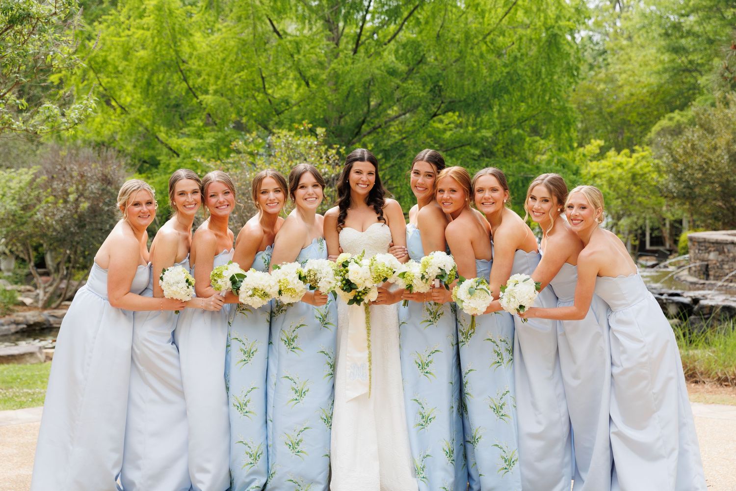 Bride and bridesmaids in light blue floral gowns holding white bouquets pose together outdoors at a wedding.