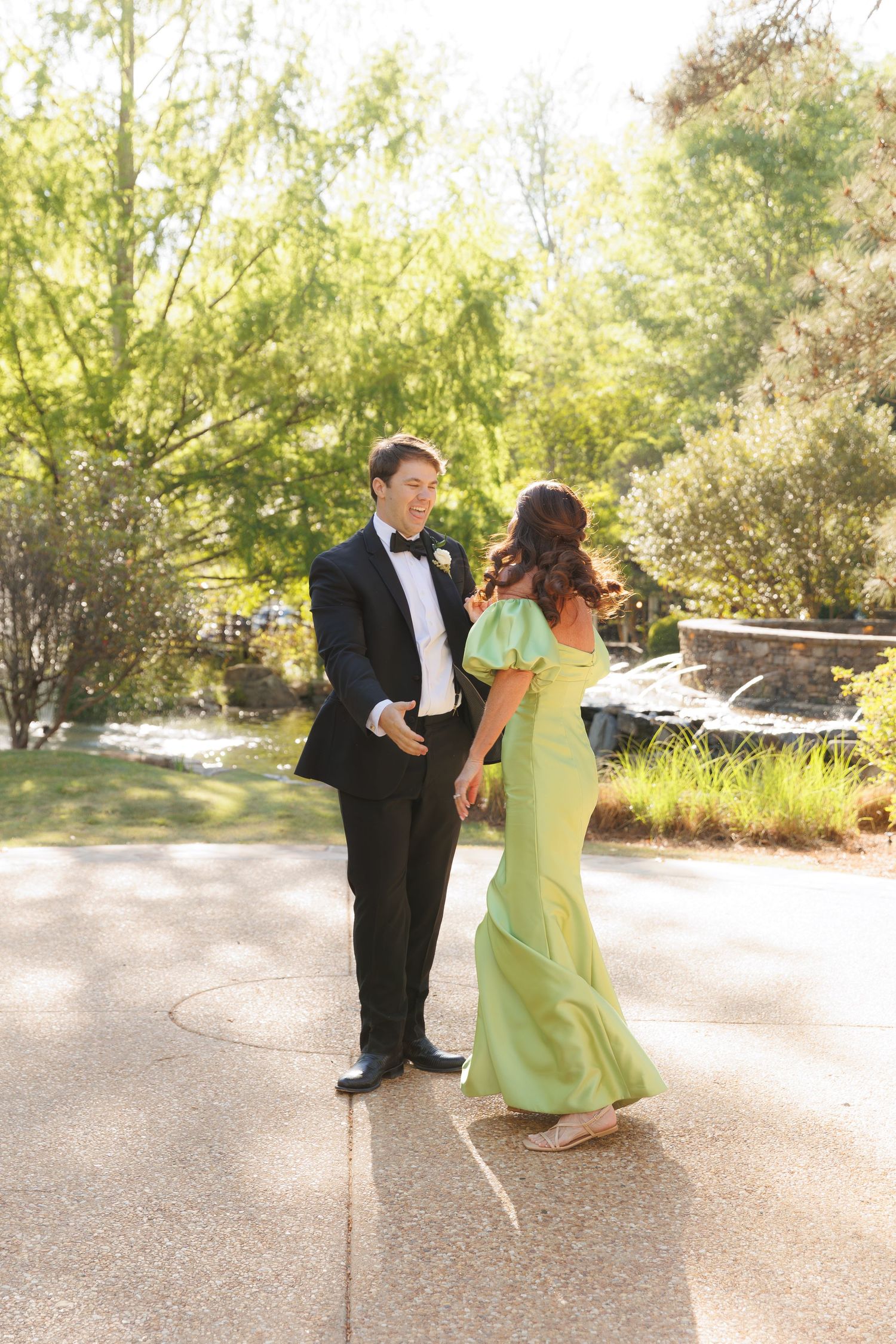 Man in black tuxedo and woman in mint green off-shoulder gown dancing together in a sunlit garden.