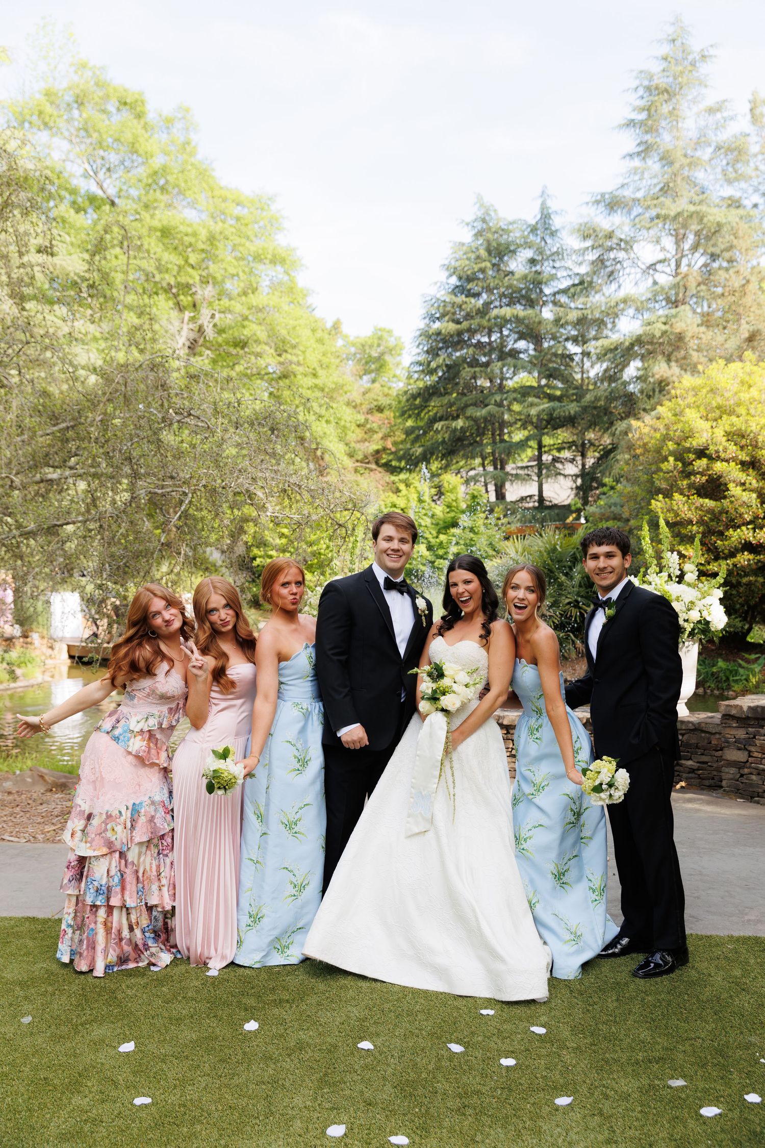 Wedding party poses outdoors, bride in white gown with bridesmaids in blue floral dresses and groomsmen in tuxedos.